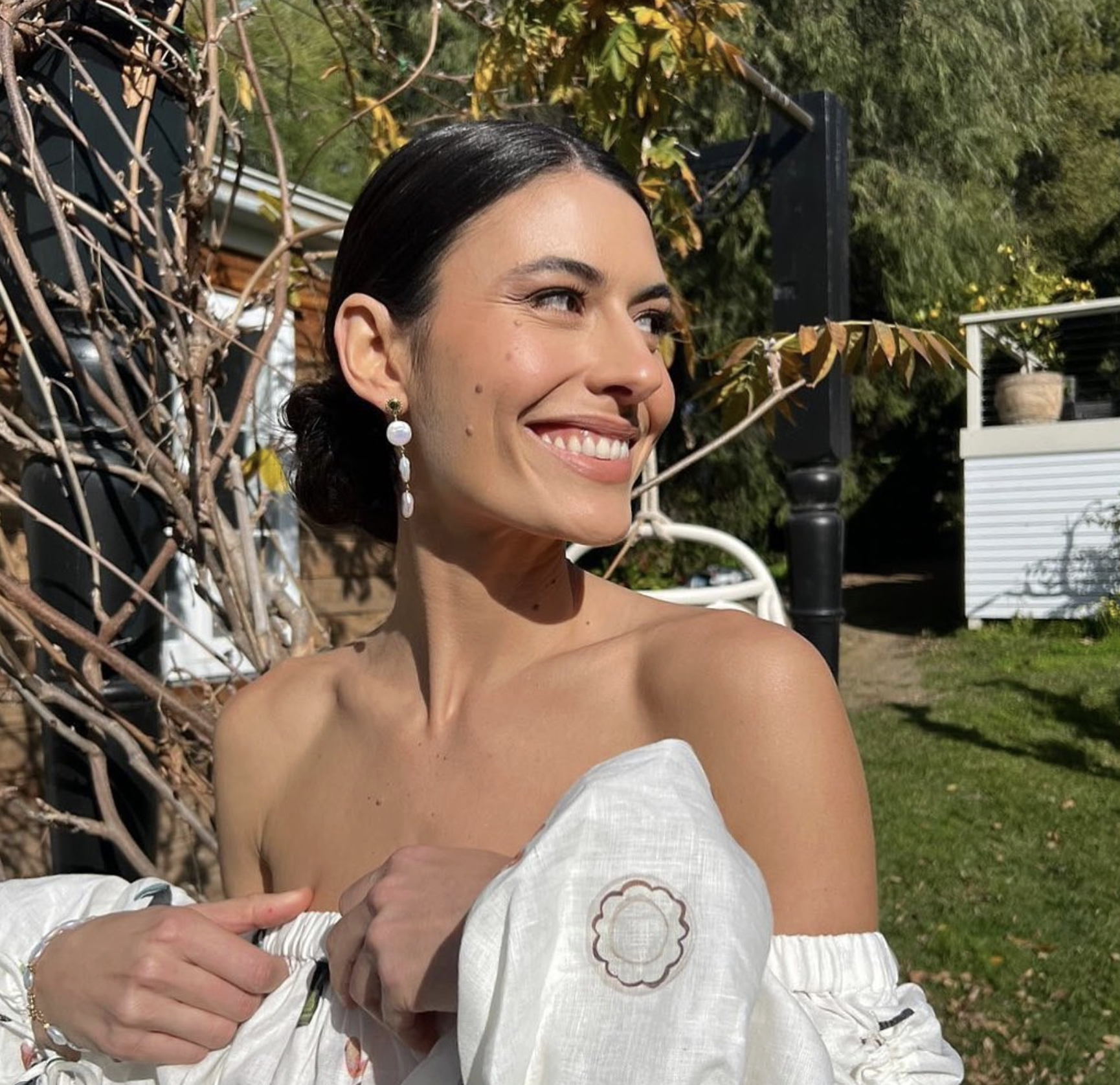 A woman with dark hair in a bun, smiling and looking to the side, outdoors with trees and a white building in the background.