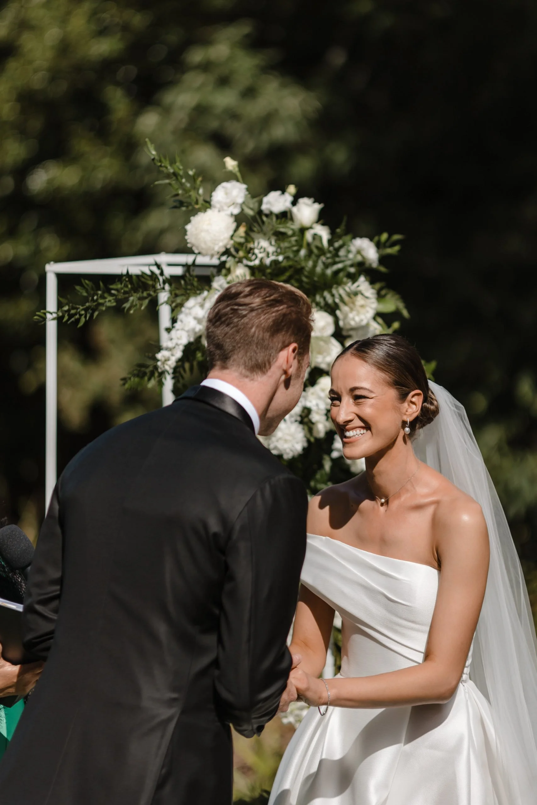 A bride and groom holding hands during their wedding ceremony, smiling at each other, with a floral backdrop of white flowers and greenery.