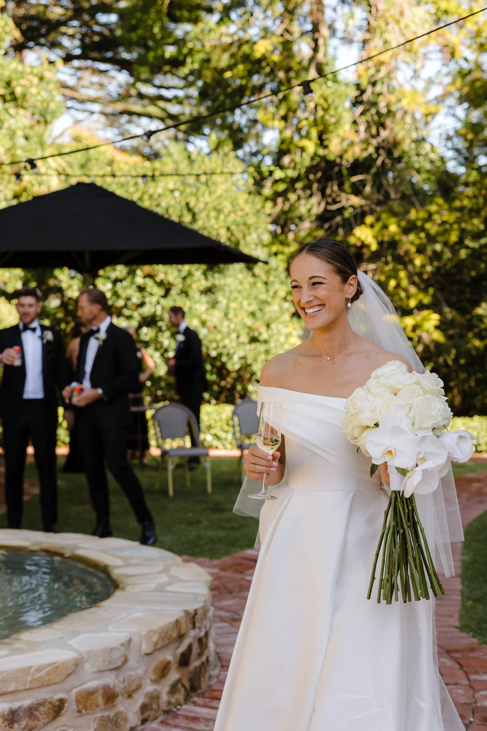 A bride in a white wedding dress holding a glass of champagne and a bouquet of white flowers, smiling at an outdoor wedding reception with guests in the background.