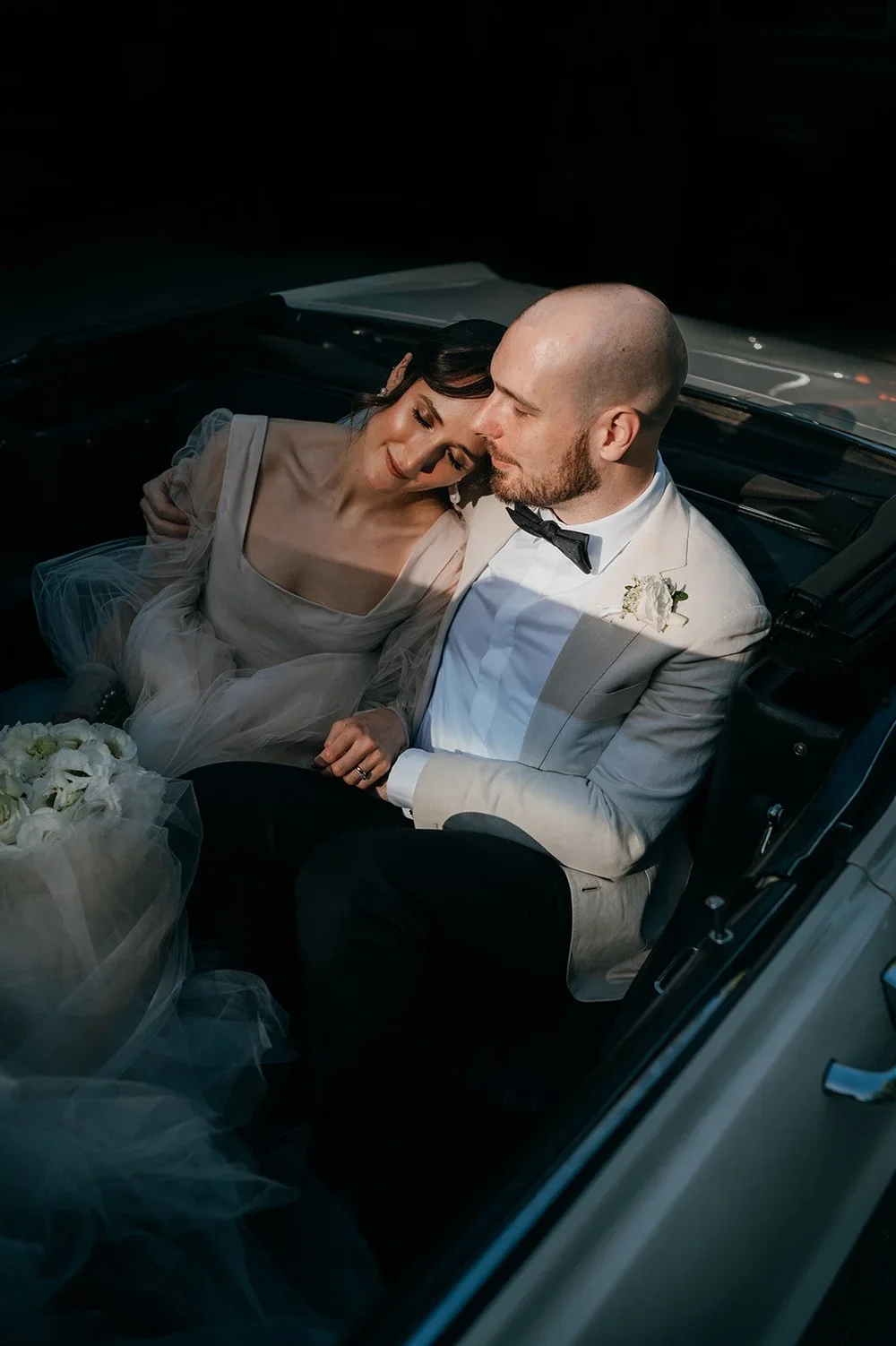 Bride and groom sitting inside a car, sharing a tender moment on their wedding day, with the bride leaning her head on the groom's forehead.