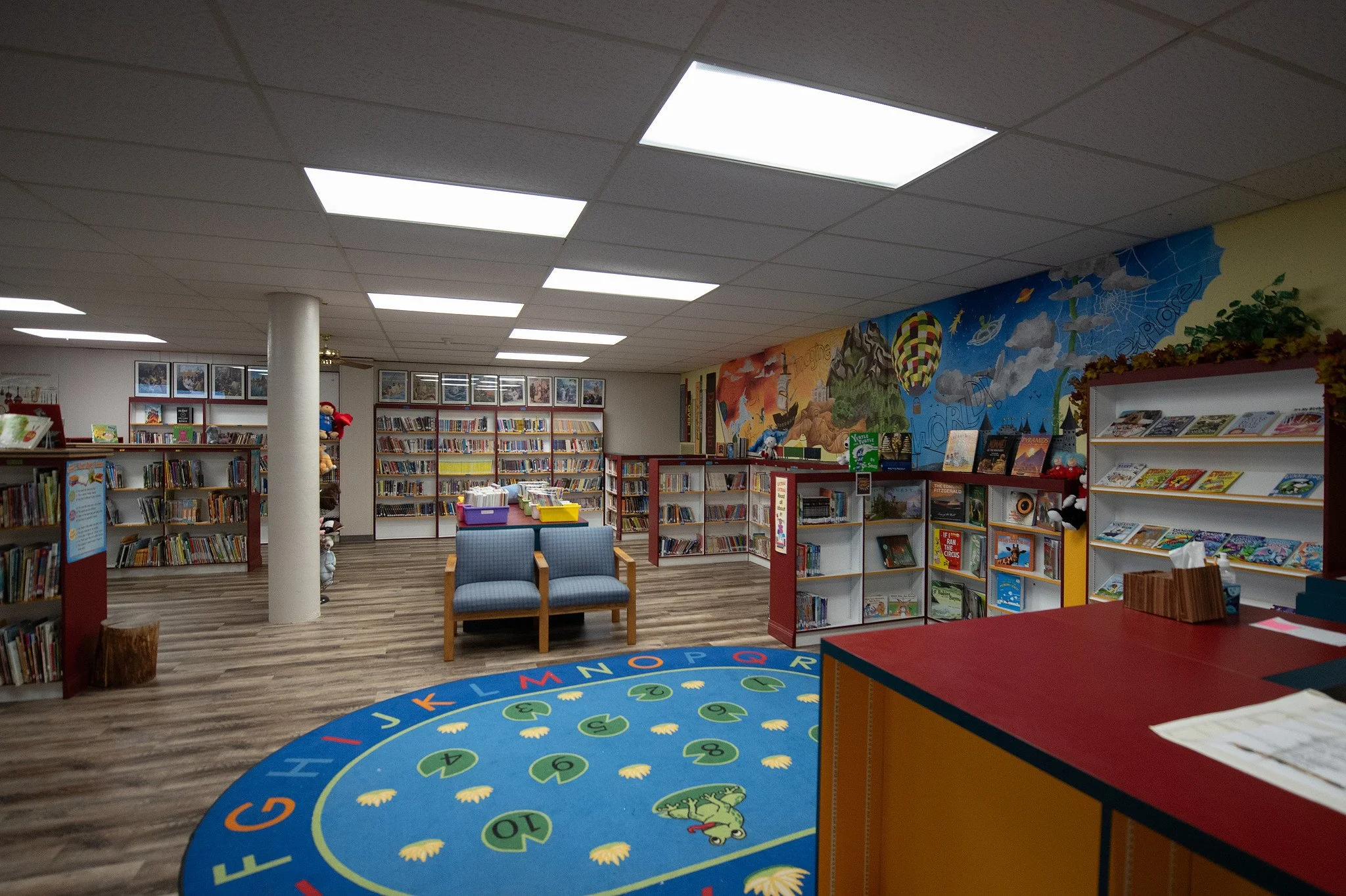 Inside a children's library with colorful mural on the wall, bookshelves filled with children's books, carpet with alphabet and numbers, blue chairs, and bright ceiling lights.