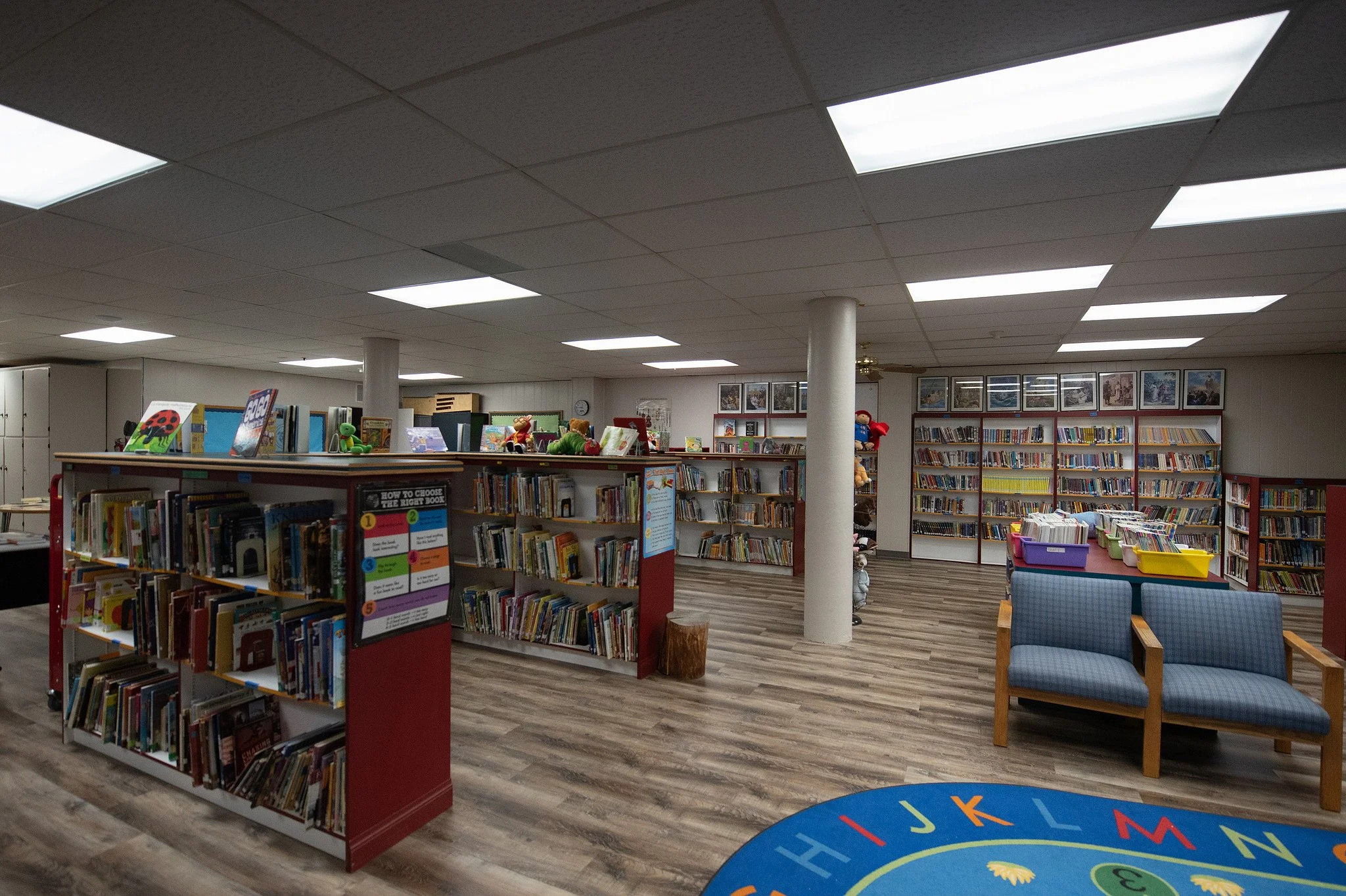 Interior of a small library with bookshelves filled with children's books, some plush toys on top, a seating area with blue cushioned chairs, and a colorful alphabet-themed rug on the wooden floor.