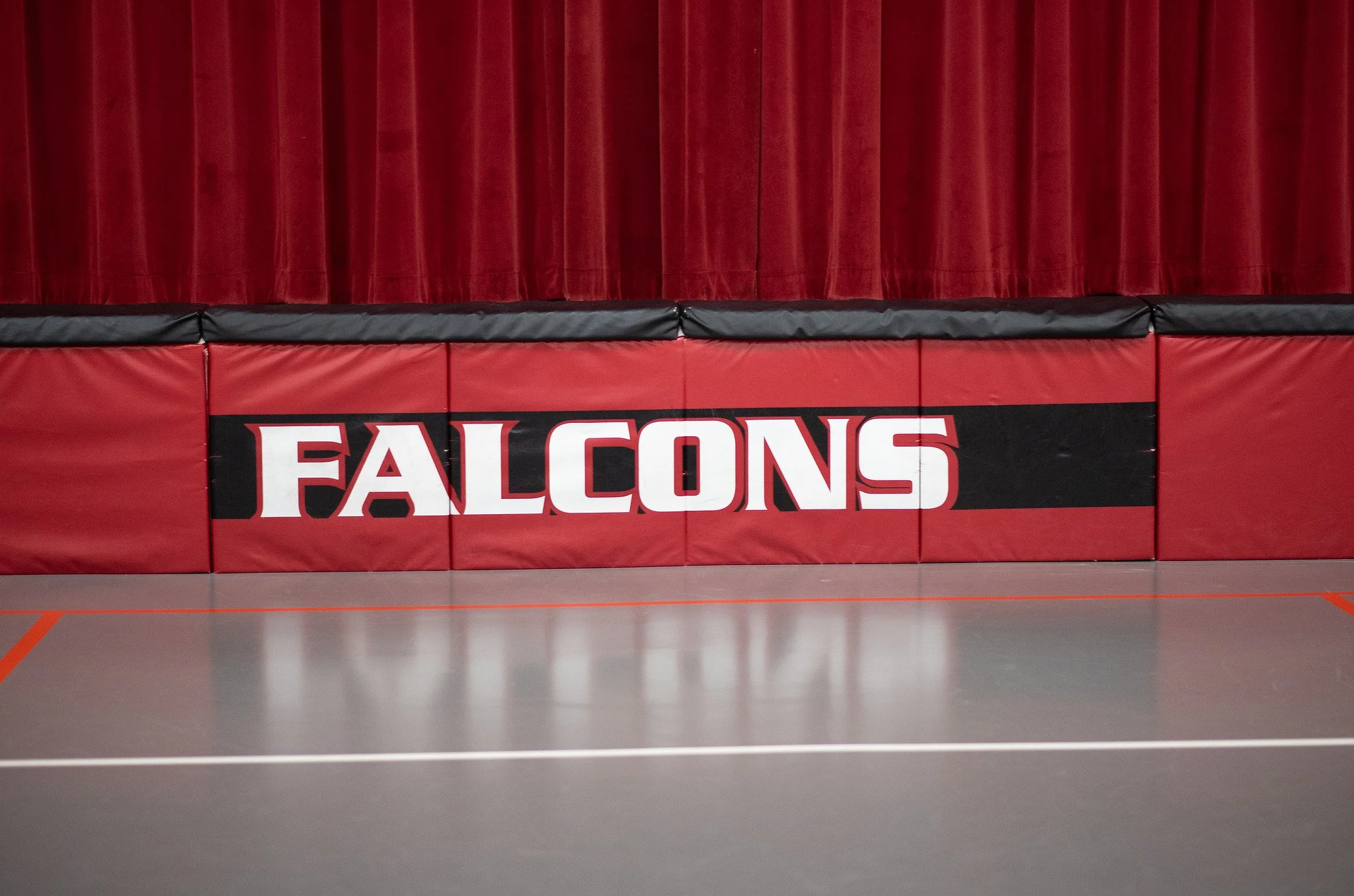 Red gymnasium wall padding with the word 'FALCONS' in large white and red letters on a black stripe, in front of red gym curtains.