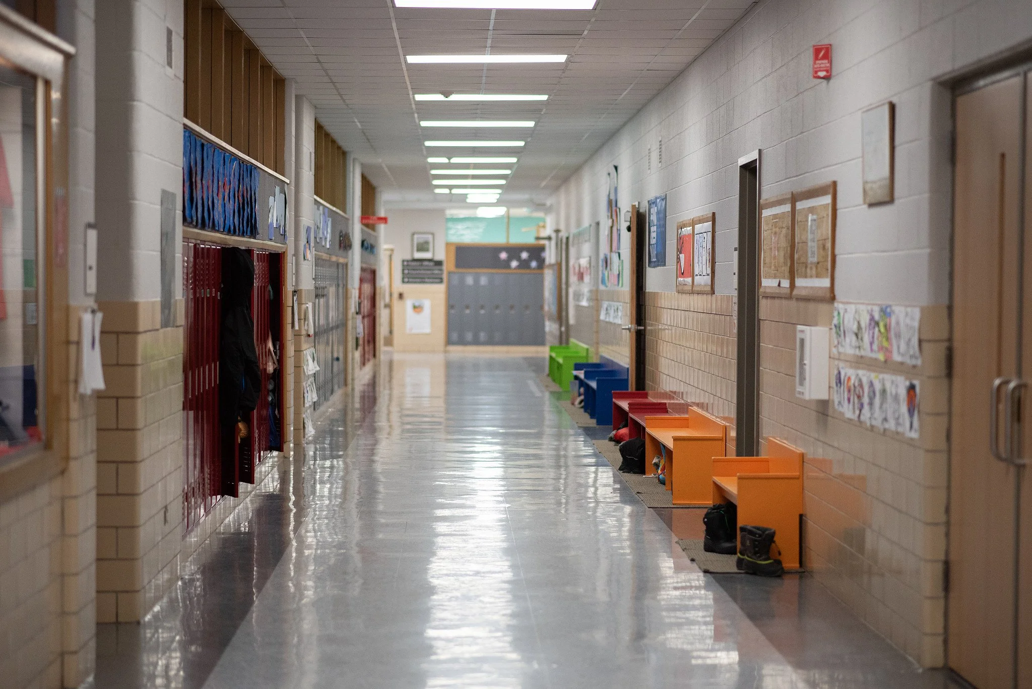 Empty school hallway with lockers on the left and cubbies on the right, decorated with artwork and a fire alarm mounted on the wall.