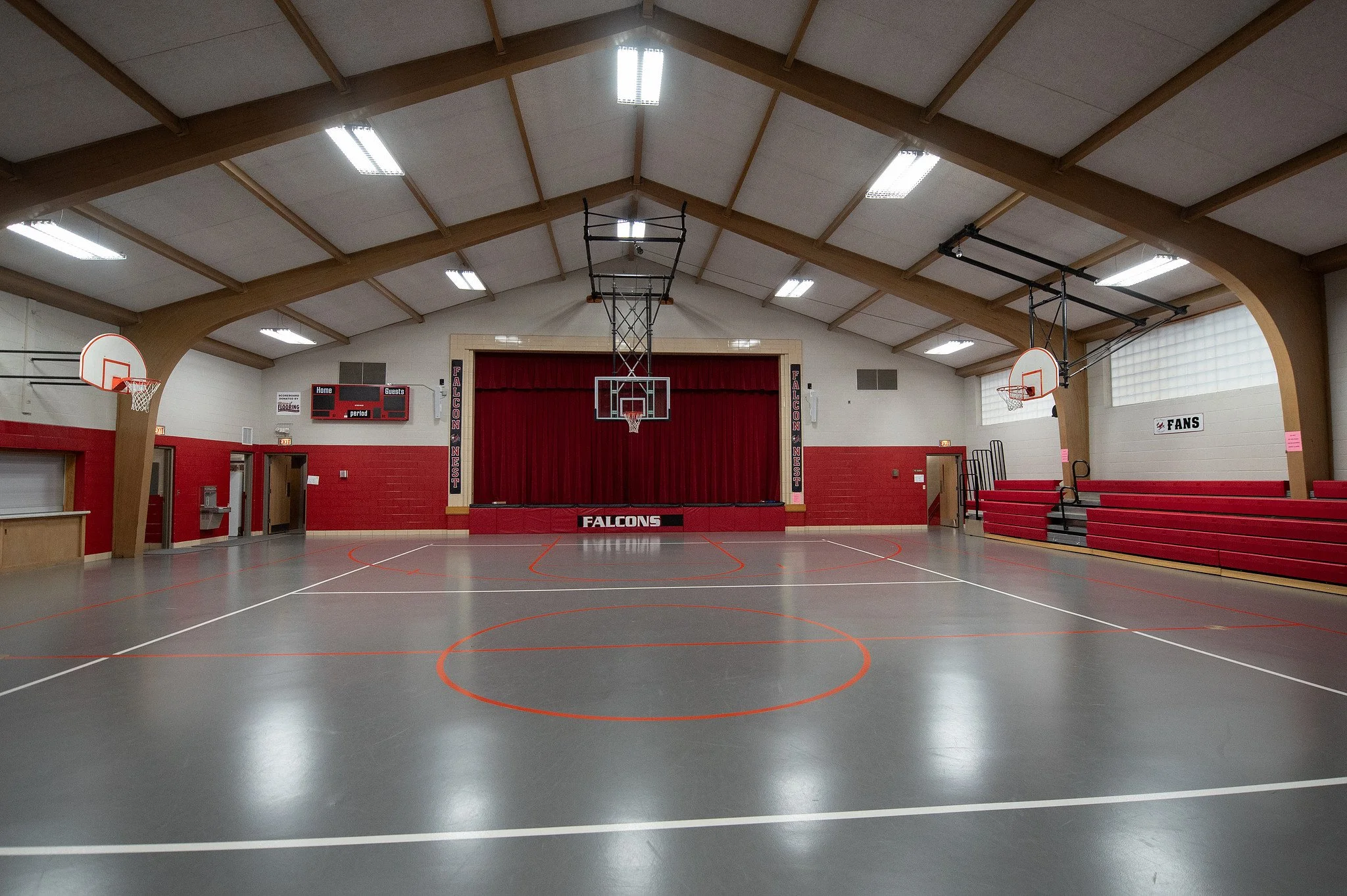 Empty indoor high school gymnasium with basketball hoops, red and gray flooring, red bleachers, and a stage with a red curtain.