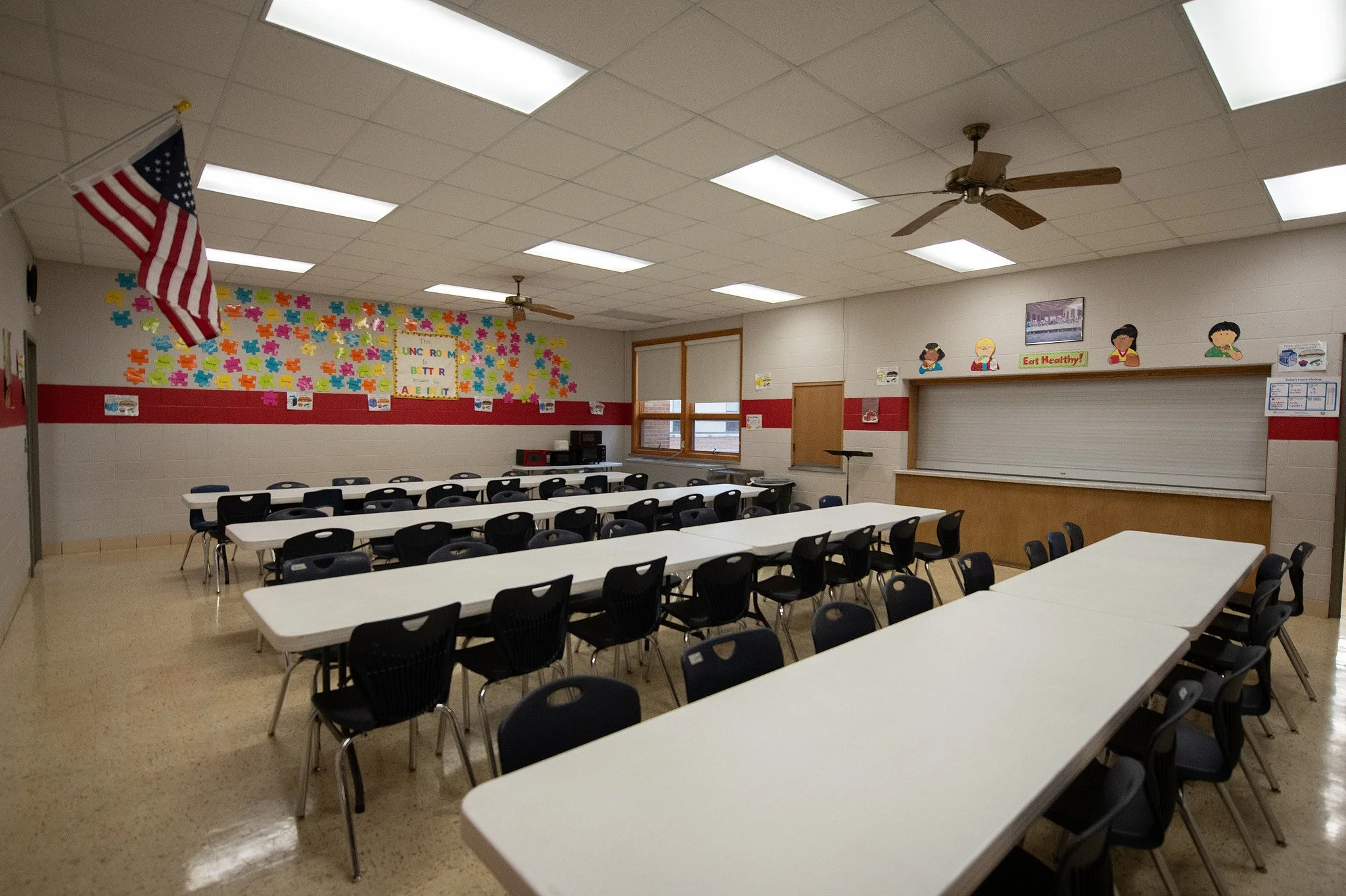 Empty classroom with white tables and black chairs, decorated with colorful puzzles on the wall, American flag, ceiling fans, and an educational display on the back wall.