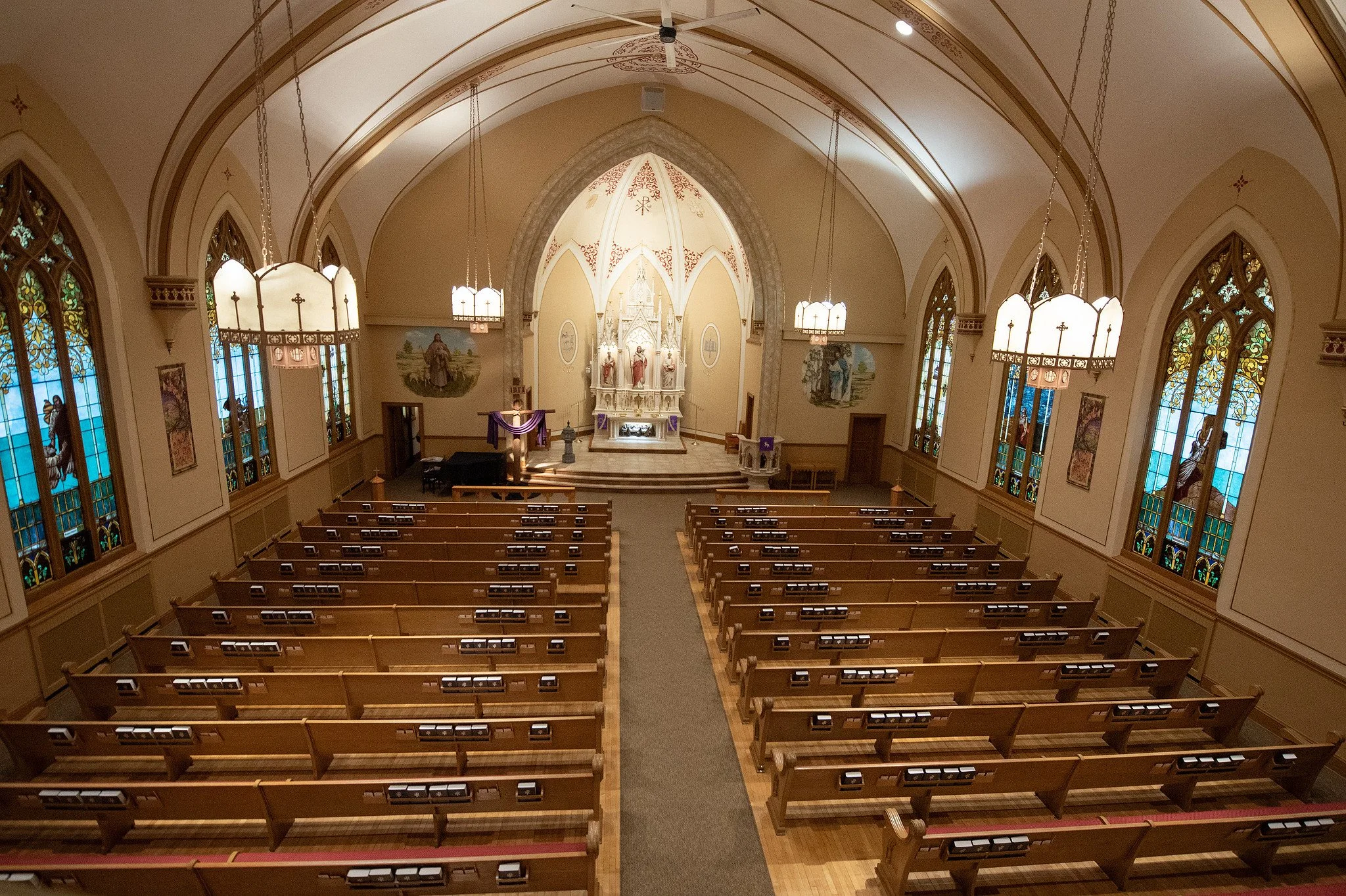 Interior of a church with wooden pews, stained glass windows, chandeliers, and altar at the front with religious statues and artwork.