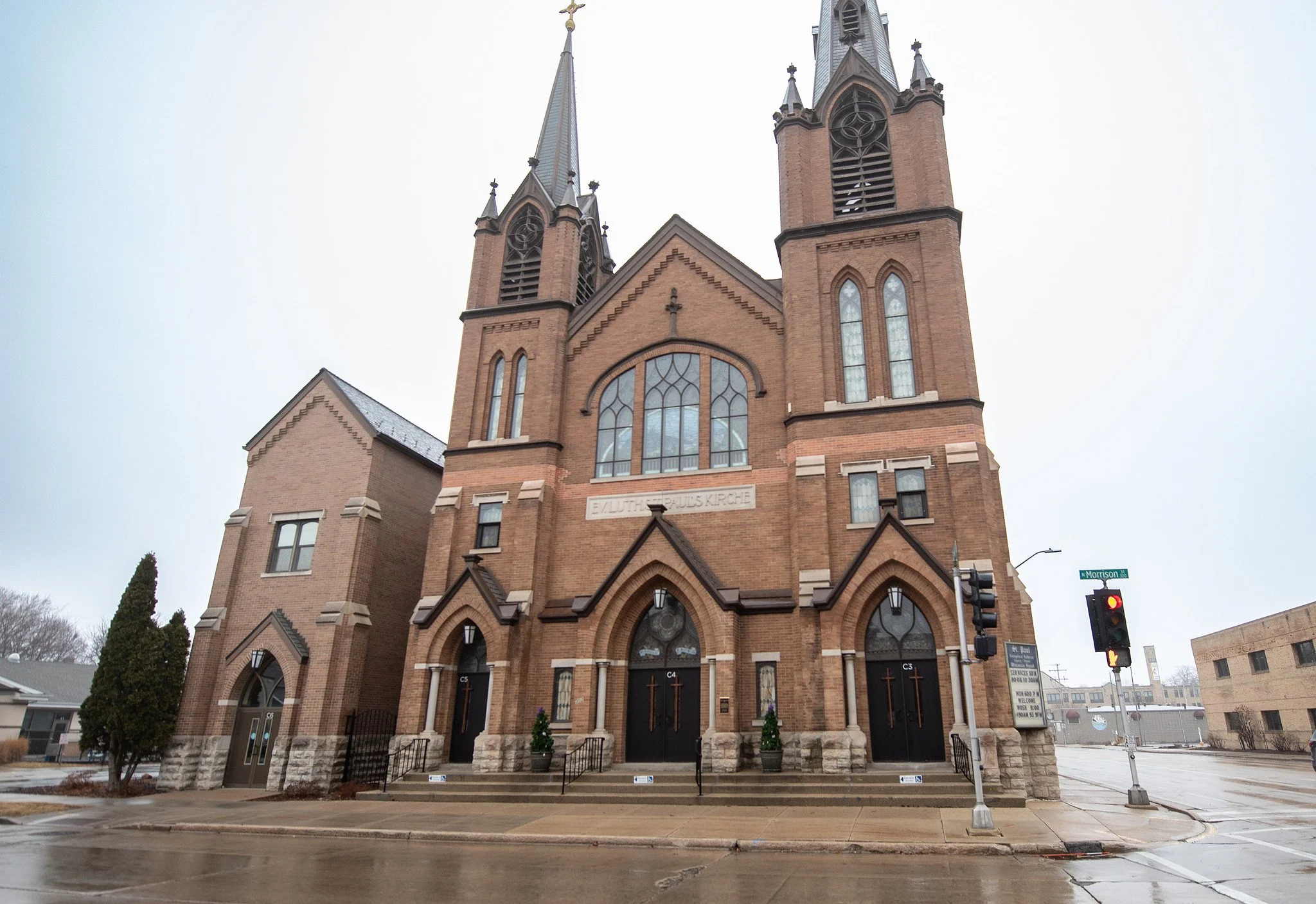 A large brick church with two tall steeples, arched windows, and stained glass, located on a street corner with traffic lights, a street sign, and a wet pavement.
