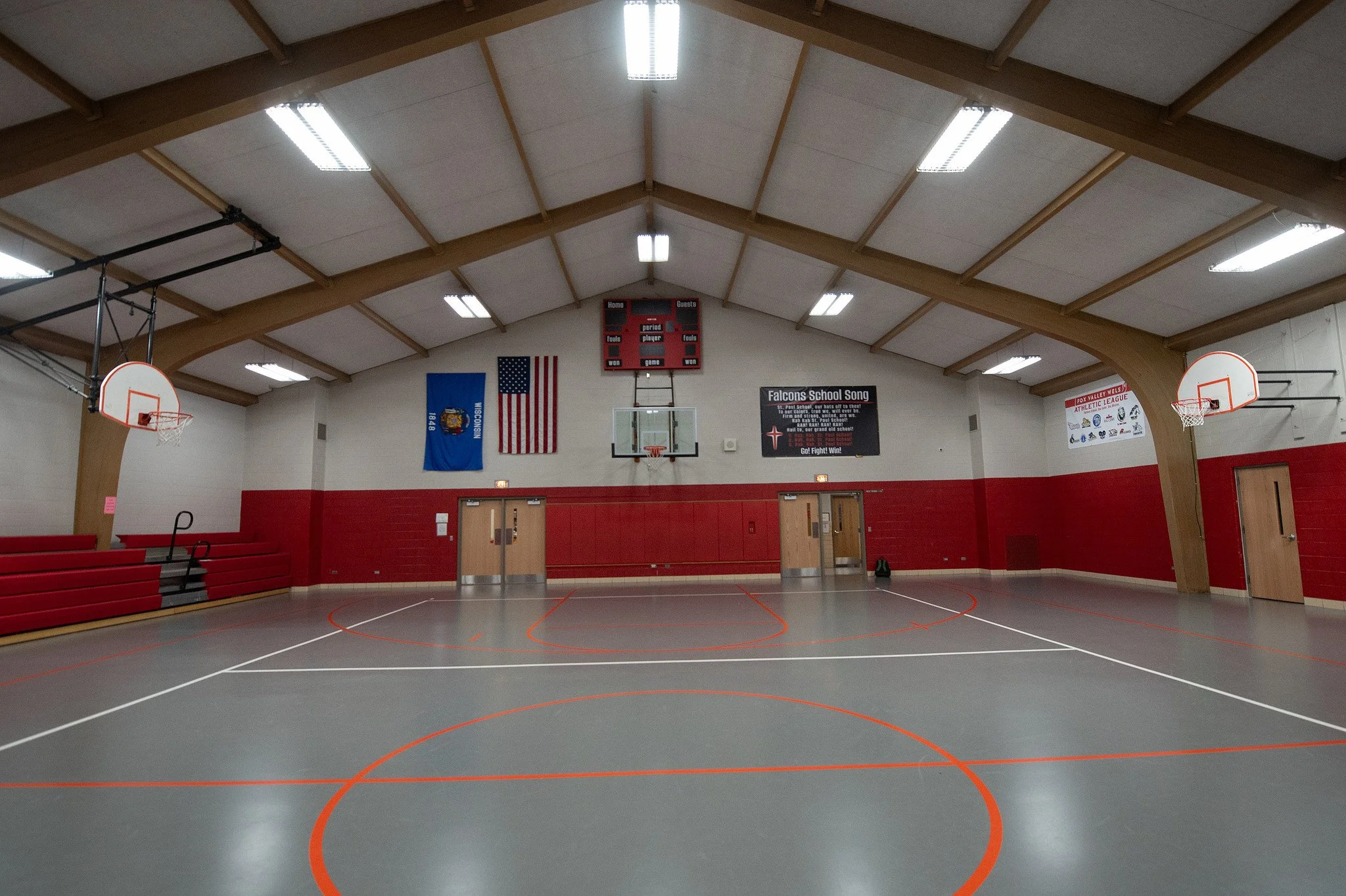 An empty indoor basketball court with red, white, and gray flooring, red walls, and a high vaulted ceiling with wooden beams. There are two basketball hoops on opposite ends and benches on one side. Flags and banners are displayed on the walls, inclu