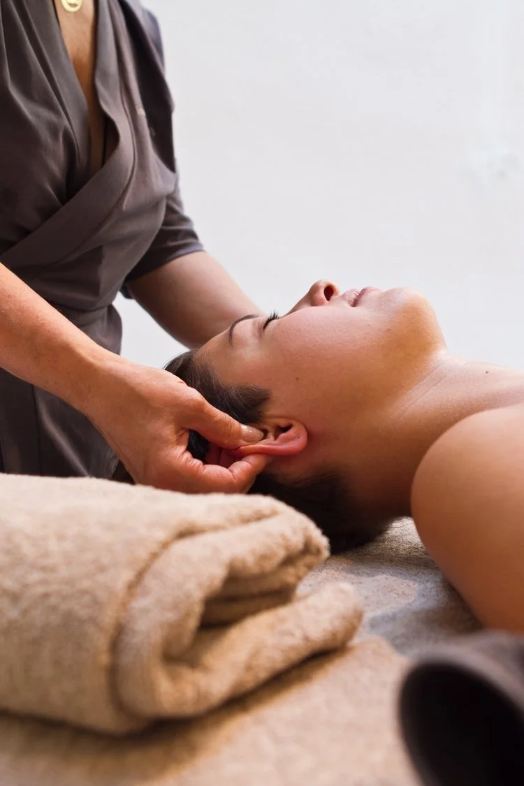 A woman receiving an ear massage or treatment from a professional in a spa or wellness setting, lying on her back with her eyes closed.