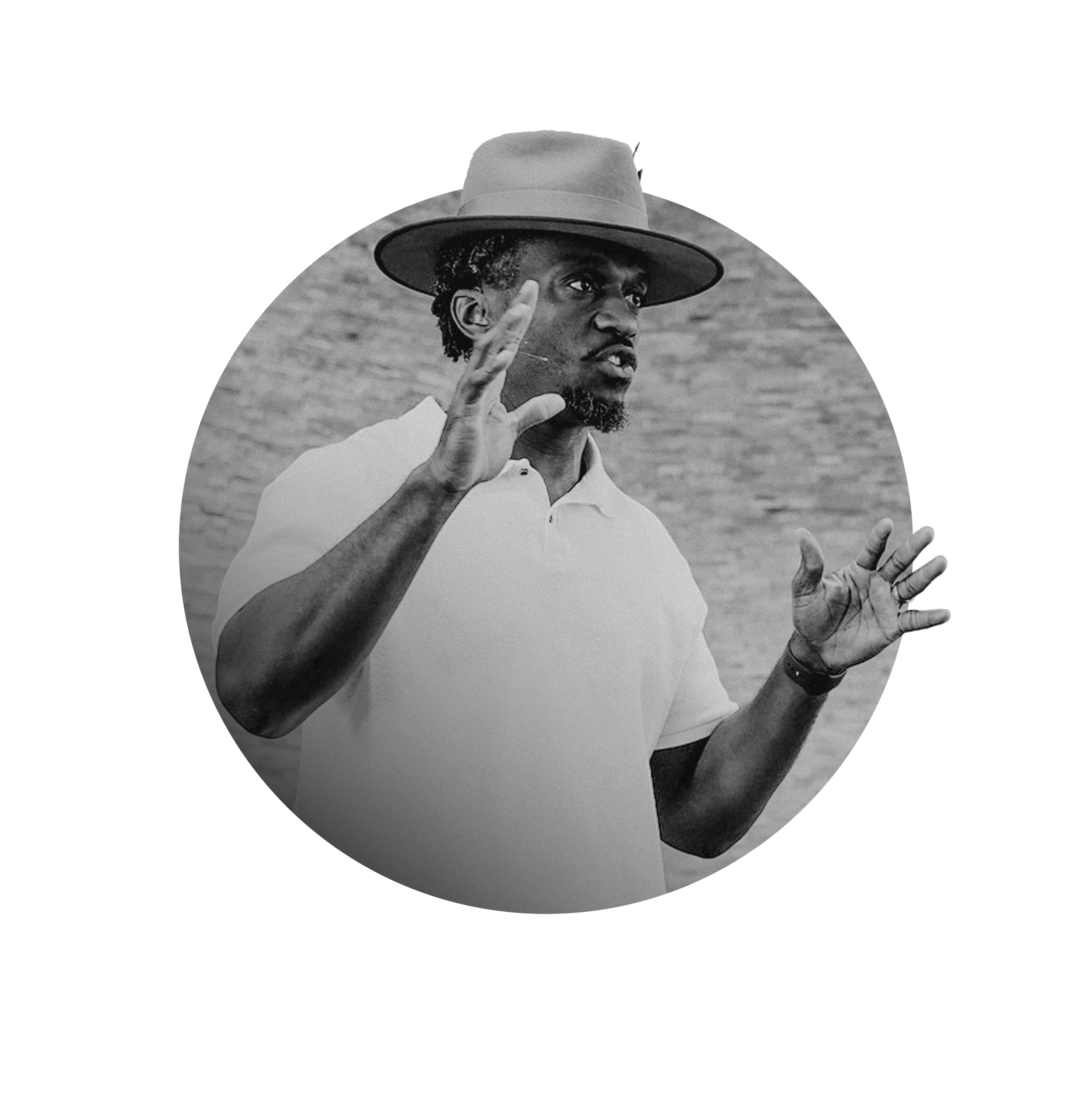 Black and white photo of Dhati Lewis, Visionary Pastor of Blueprint Church in Atlanta, Georgia, wearing a hat and a white polo shirt, gesturing with his hands as if speaking or explaining something, standing outdoors against a brick wall background.