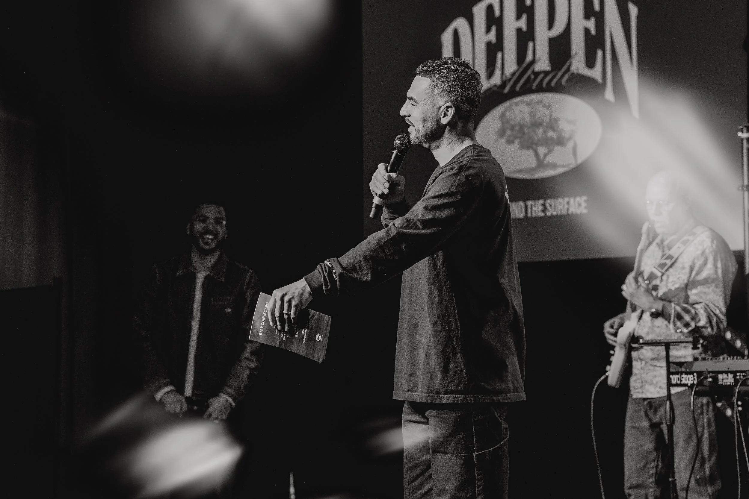 A man with a microphone on stage, holding a booklet, with two musicians in the background, one playing guitar and the other on keyboard. There is a large screen behind them displaying the words "Deepen" and other text.