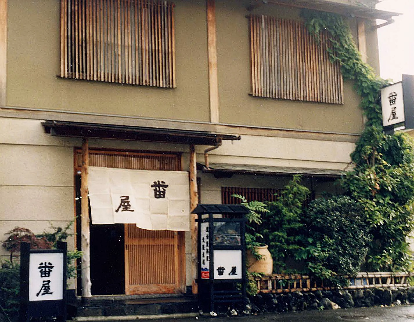 伝統的な日本の飲食店の外観。木製の入口扉と布製の暖簾に黒字で書かれた日本語の文字。周囲には緑の植物と看板が配置されている。