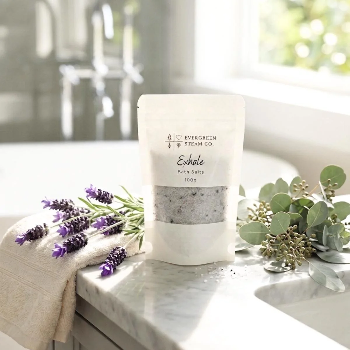 A packet of Evergreen Steam Co. Exhale bath salts on a white marble countertop, surrounded by sprigs of lavender and eucalyptus, with a beige towel nearby, in a bright, airy bathroom with sunlight coming through a window.