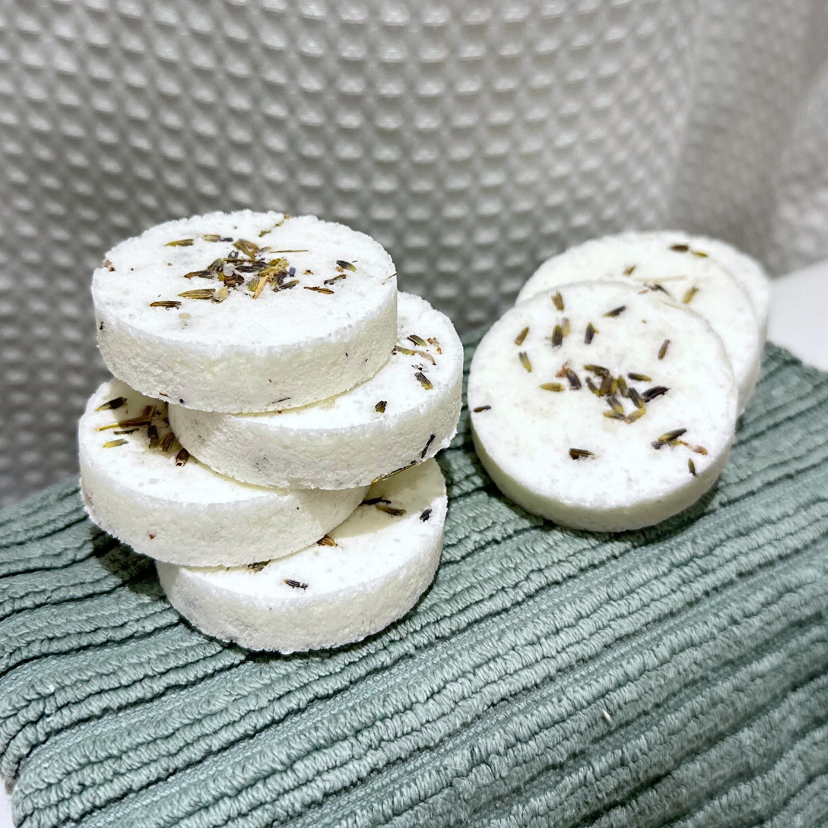Six shower steamers with dried lavender petals on top, placed on a textured green surface with a gray textured background.