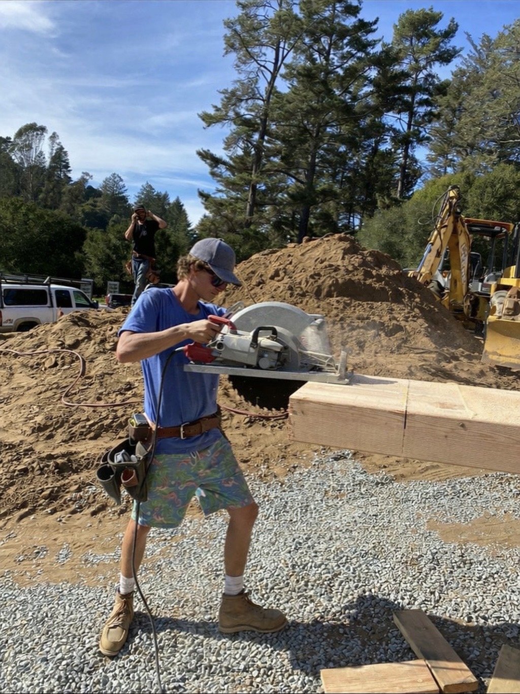 Person in blue shirt and shorts using a circular saw to cut a large wooden beam at a construction site, with a person in black and a yellow excavator in the background.