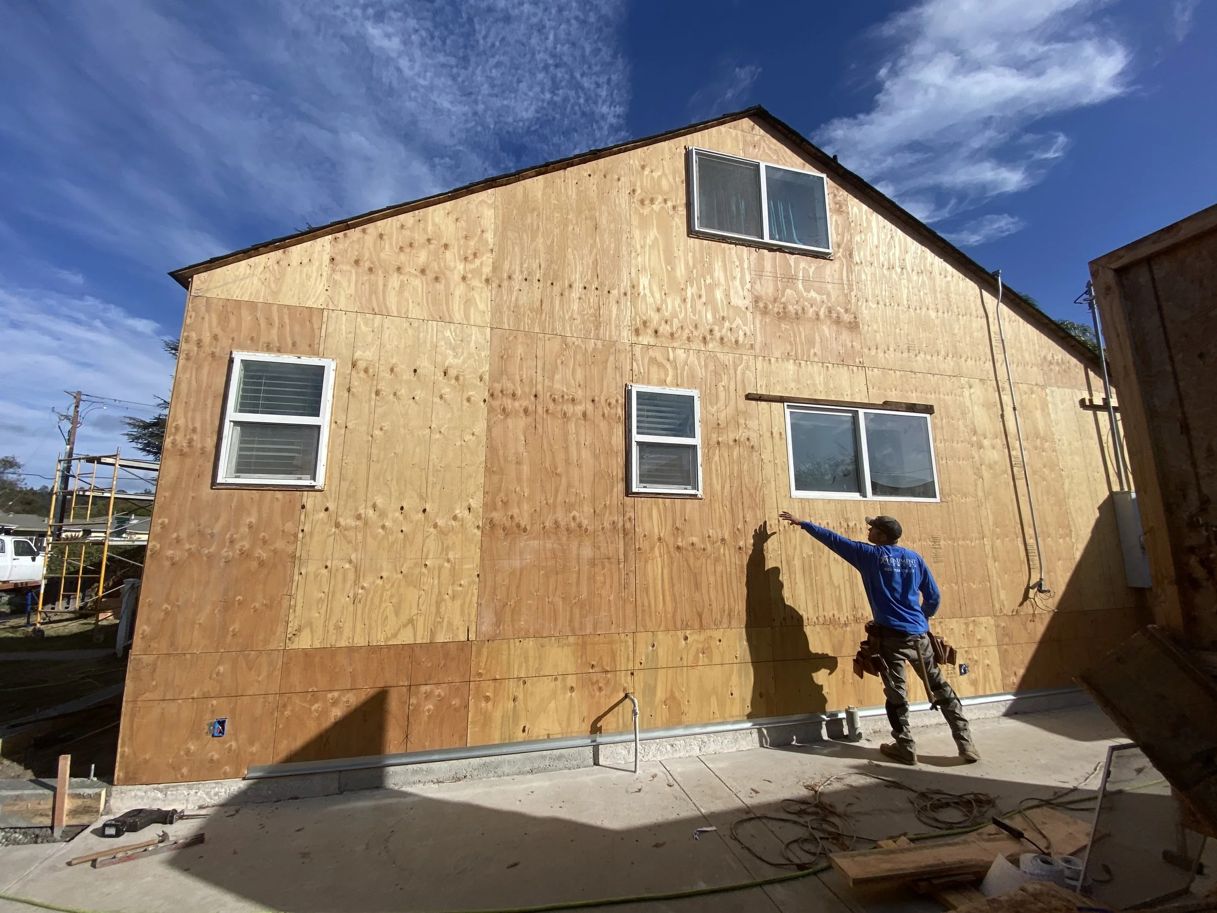Construction worker pointing at a partially built wooden house with plywood exterior, three windows, and an electrical conduit on the wall, under a blue sky with wispy clouds.