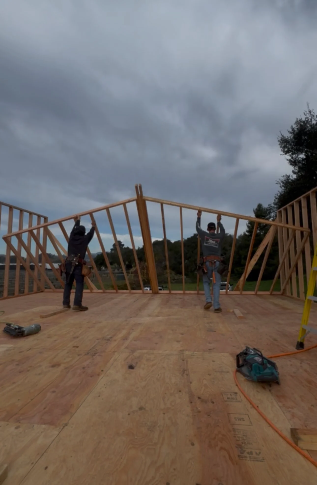 Construction workers are framing a wooden structure on a building site with a cloudy sky overhead.