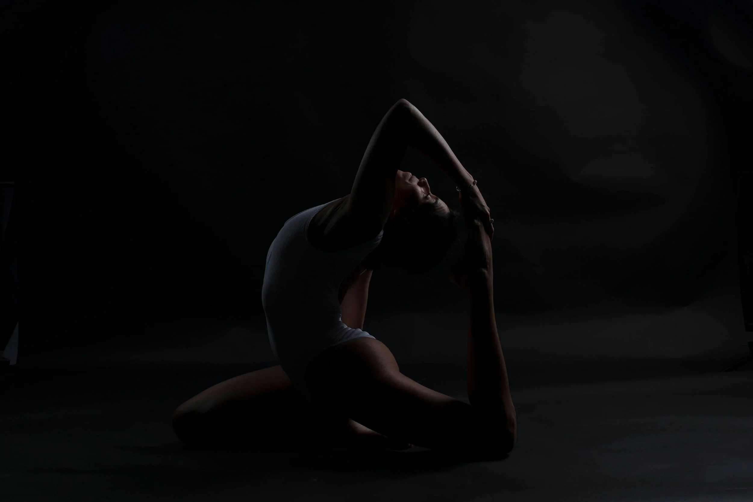 A woman performing a yoga pose on a black background, with minimal lighting highlighting her silhouette.