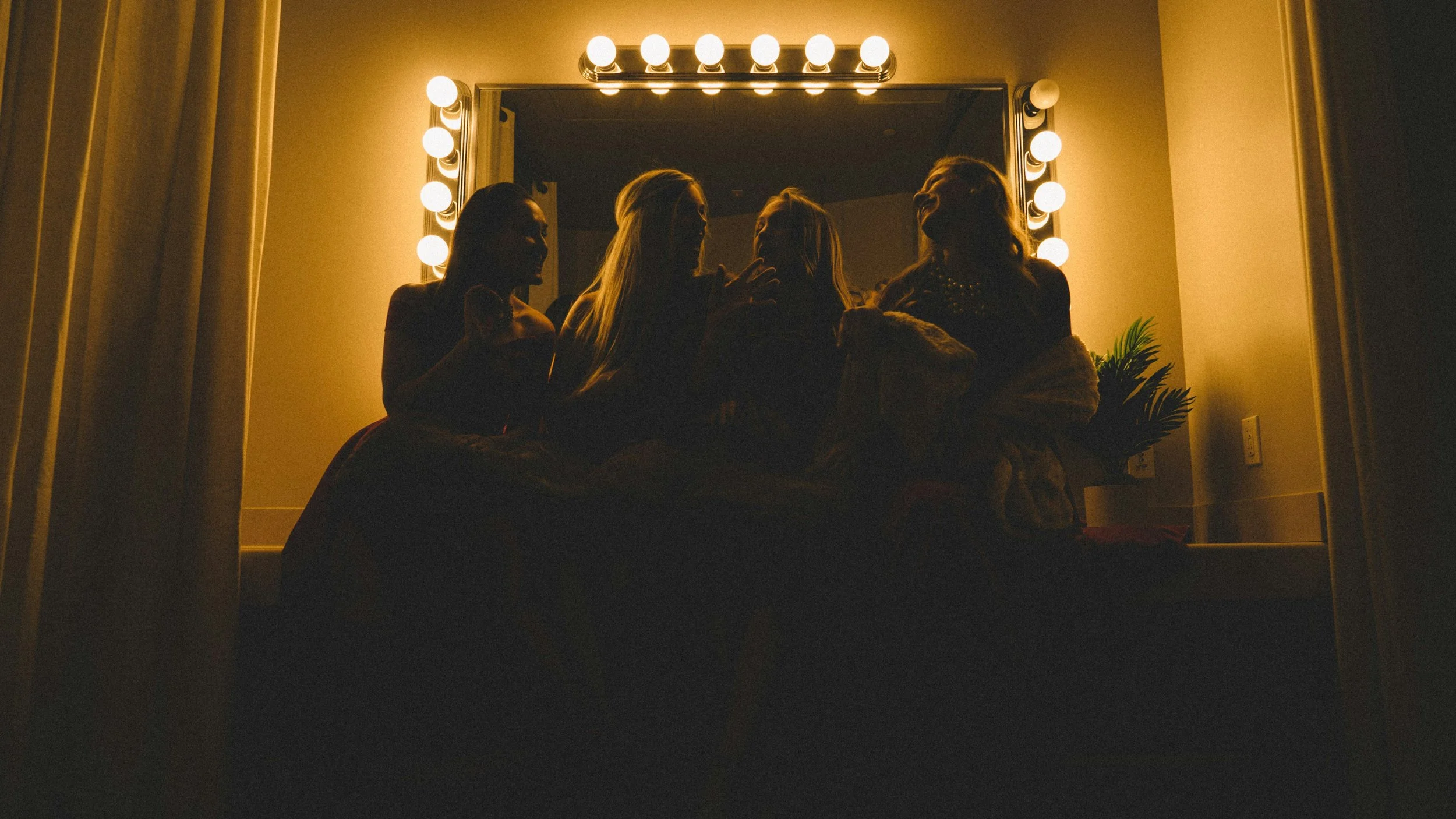 Four women sitting on a bed in front of a mirror with lights, silhouetted against the warm lighting.
