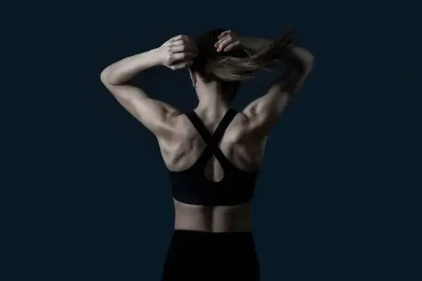 A woman with toned back muscles pulling her hair back against a dark background.
