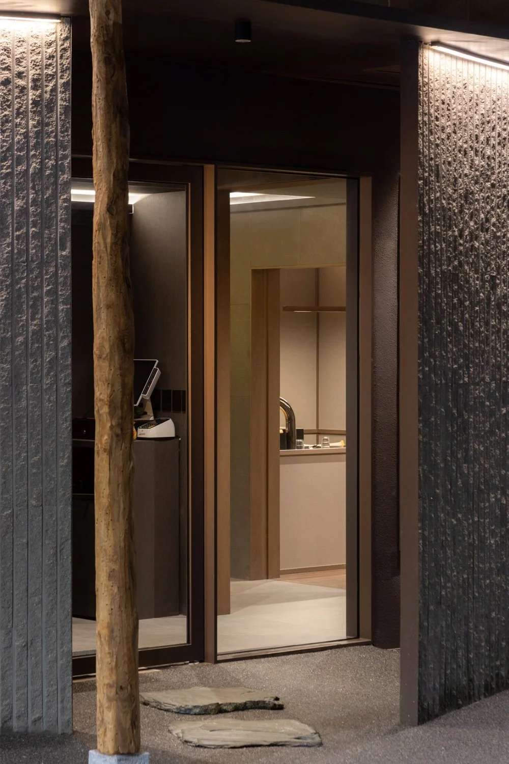Interior view of a modern room with dark walls, textured panels, and a large wooden pole, partially open sliding door revealing a kitchen with a sink.
