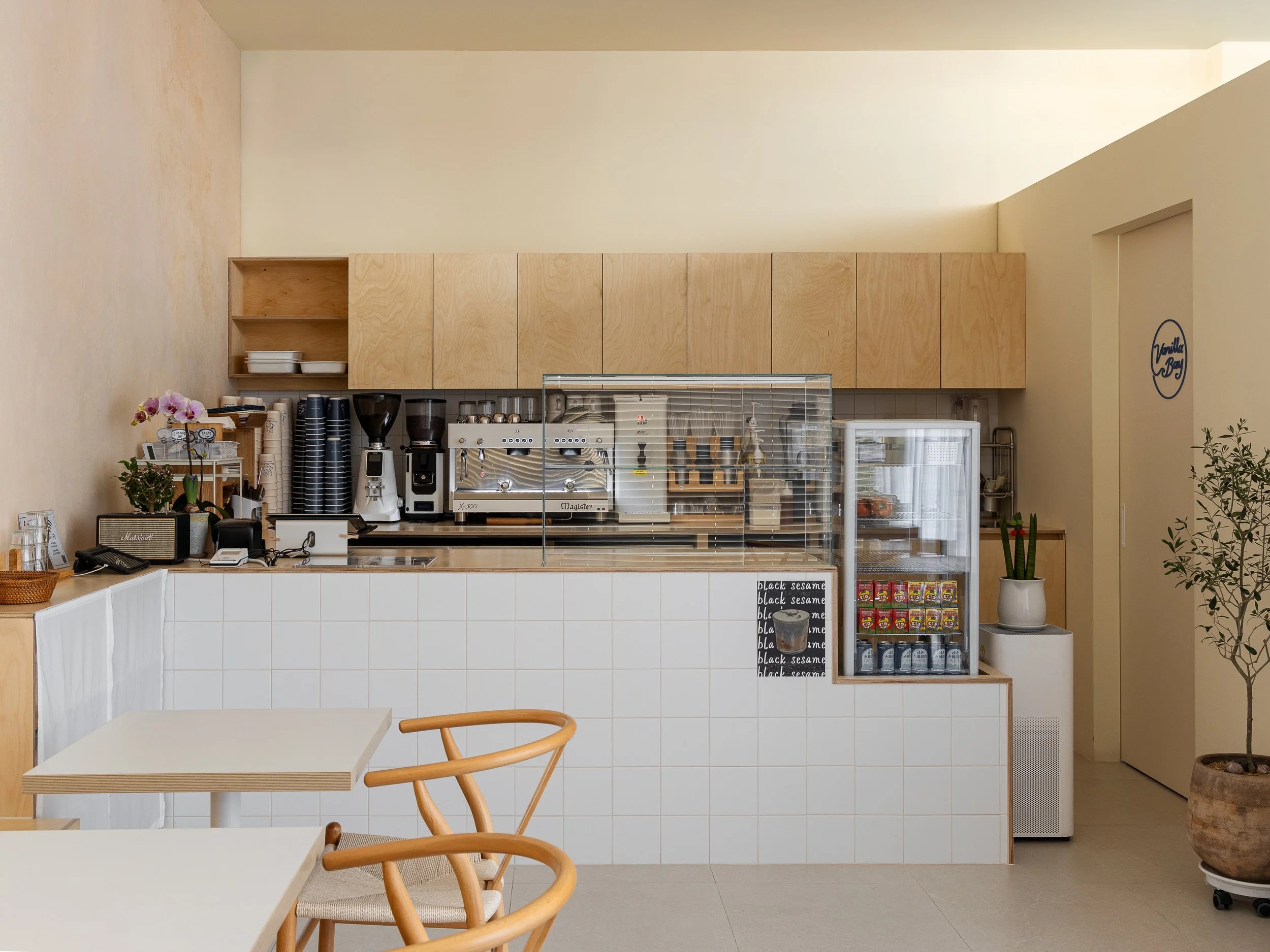 A minimalist coffee shop with a counter displaying coffee machines, cups, and snacks, and seating with wooden chairs and white tables.