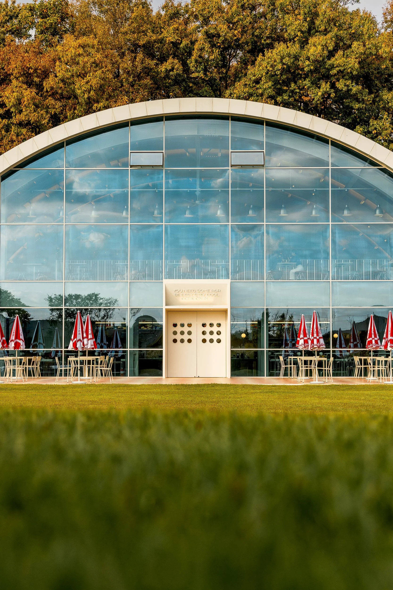 A modern building with a large, curved glass facade, red and white patio umbrellas, and outdoor seating on a grassy area, set against a background of trees with autumn foliage.
