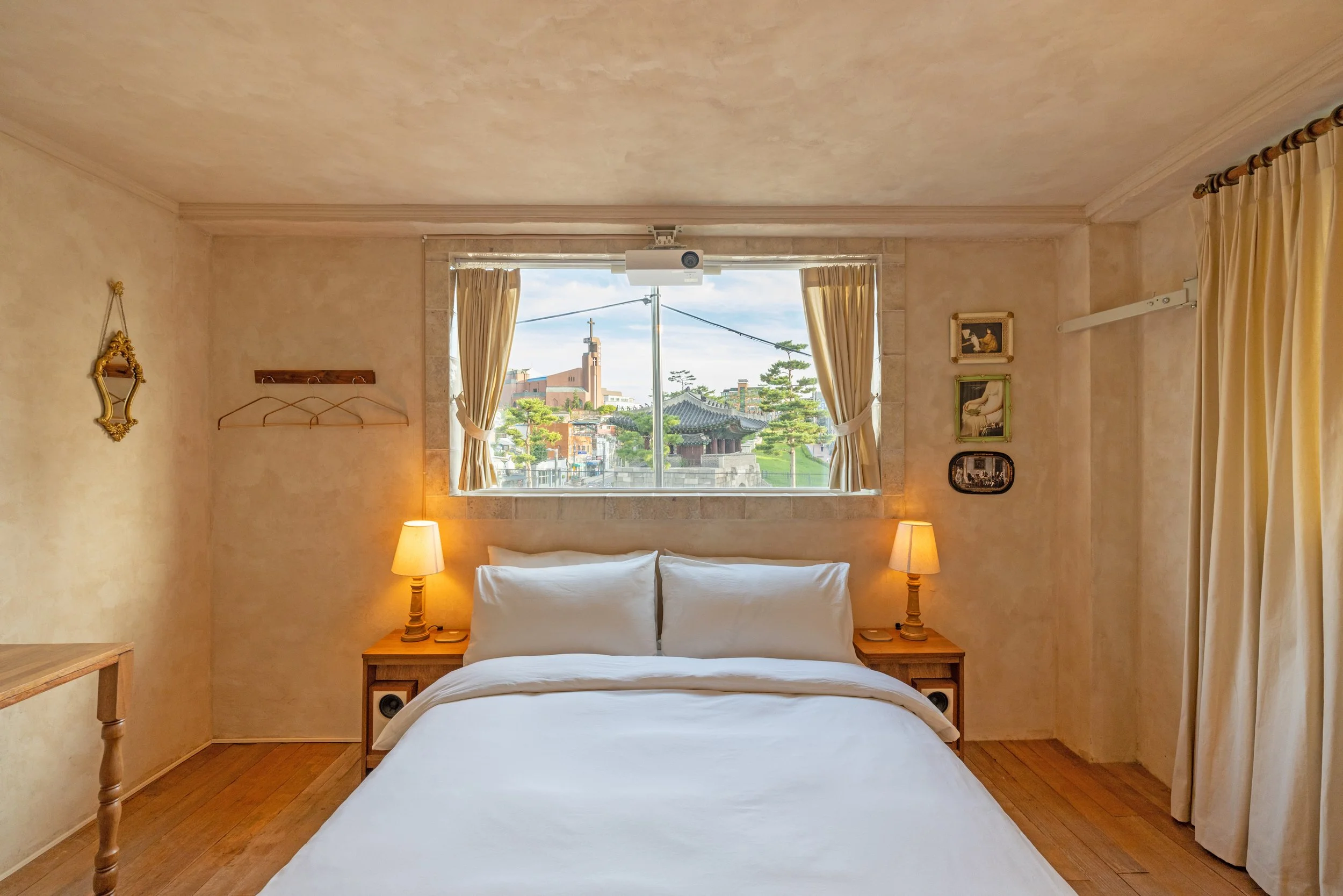 Cozy bedroom with a large window, a white bed with pillows, wooden bedside tables with lamps, and light-colored curtains.