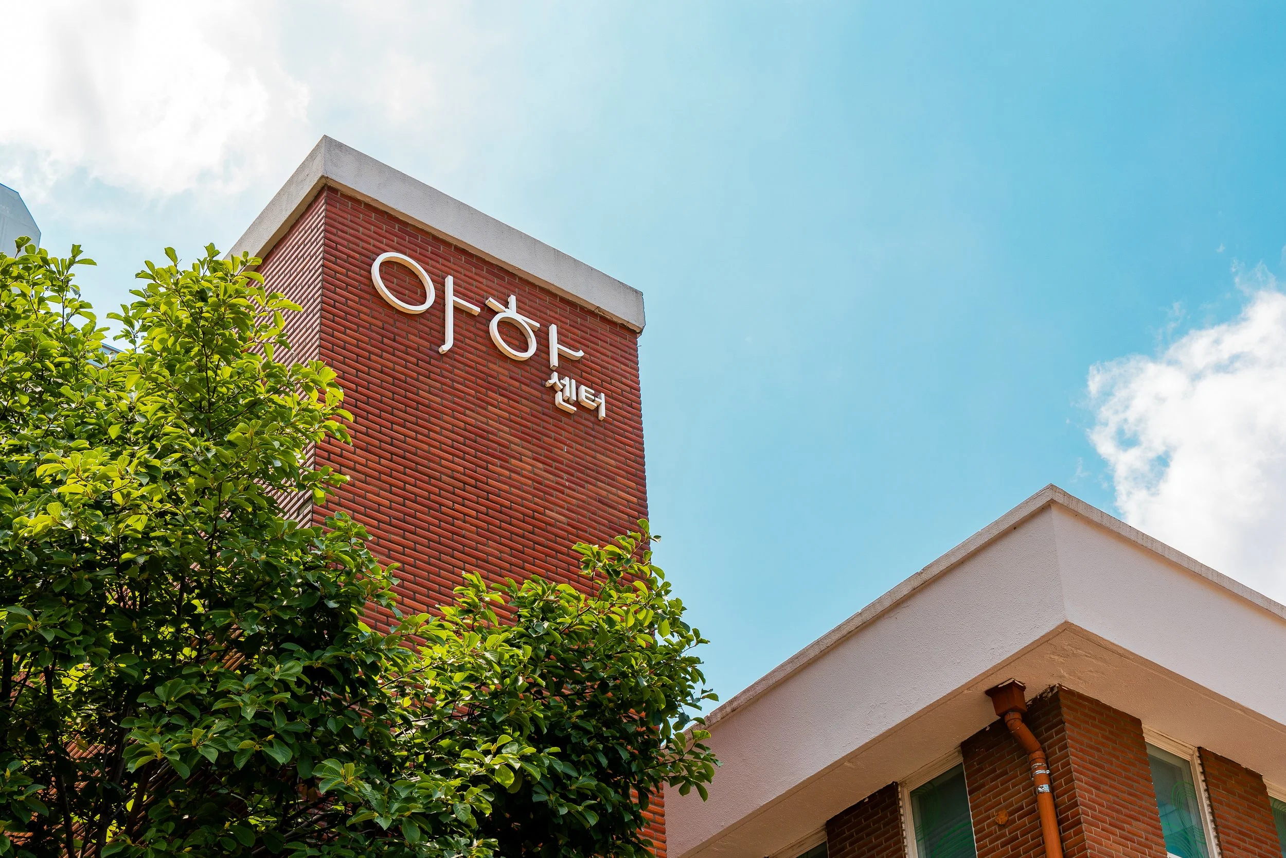 A red brick building with white lettering on the side reading '아포센터'. A leafy green tree is in the foreground, and the sky is blue with some clouds.