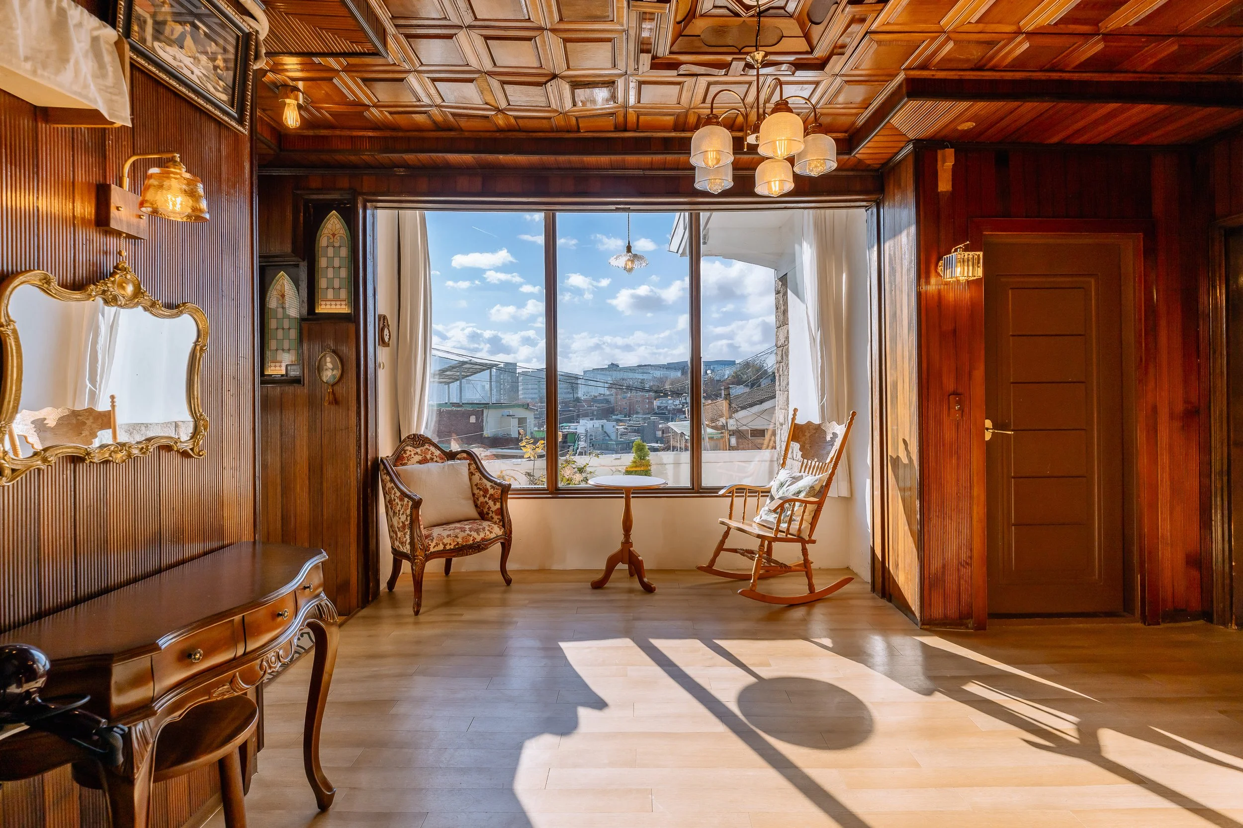 A cozy room with wooden paneling and a large window showing a sunny outdoor view with buildings and clouds. The room has vintage furniture including a floral armchair, a rocking chair, a small round table, and a wooden sideboard. There are hanging ceiling lights and ornaments on the wall.