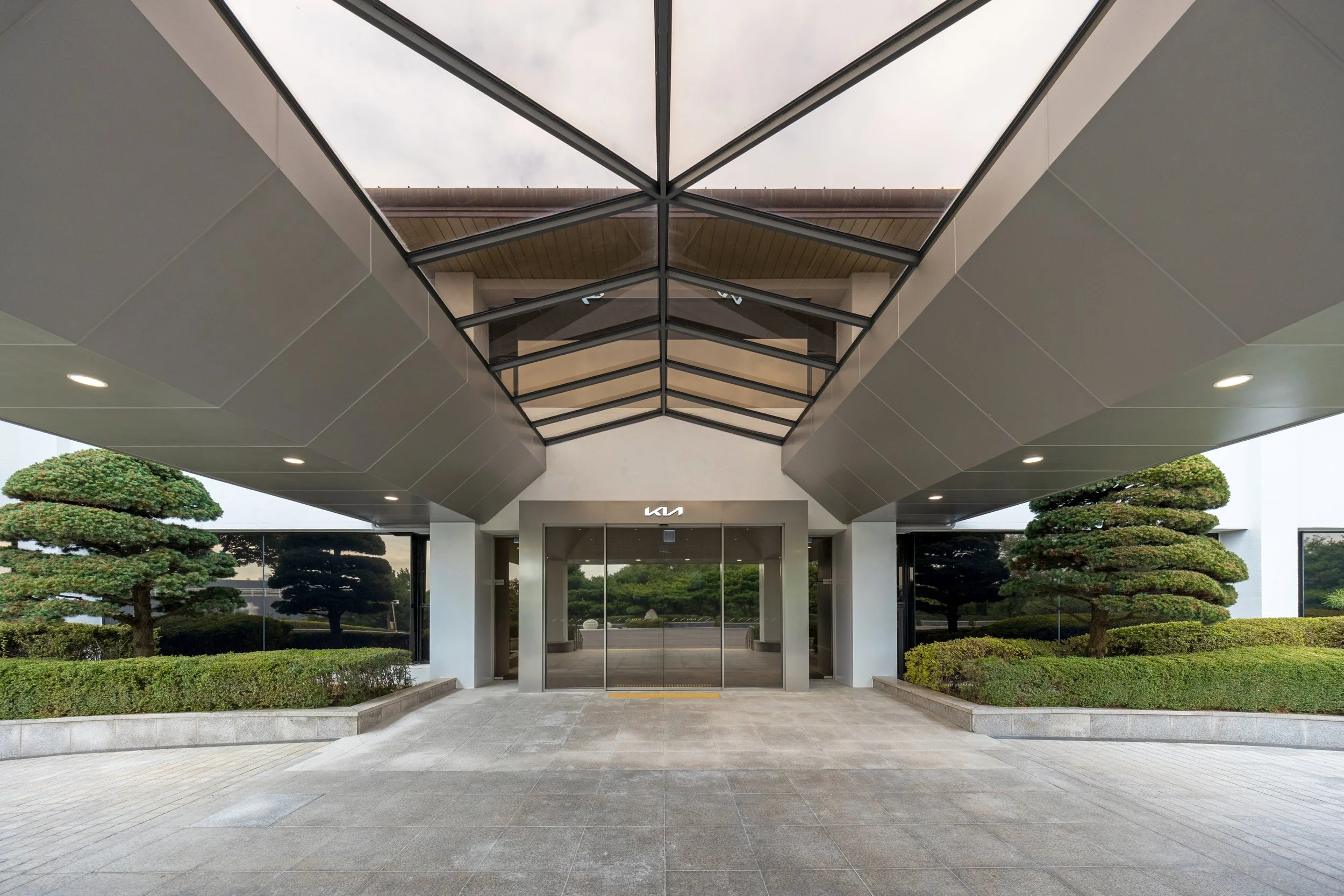 Modern building entrance with glass doors, landscaping, and a geometric glass roof overhead.