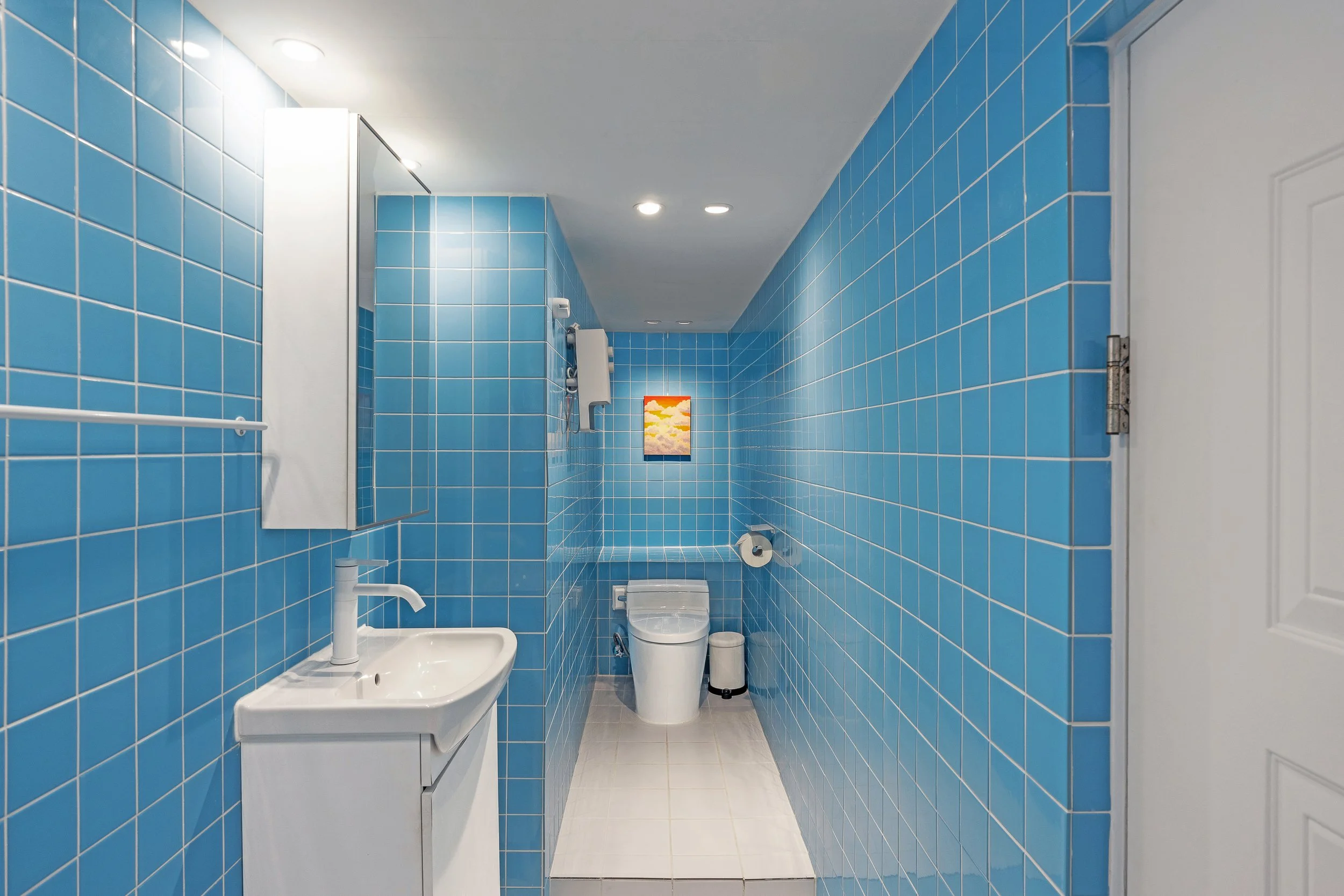 Blue tiled bathroom with white sink, mirror cabinet, toilet, toilet paper holder, and a small colorful painting on the wall.
