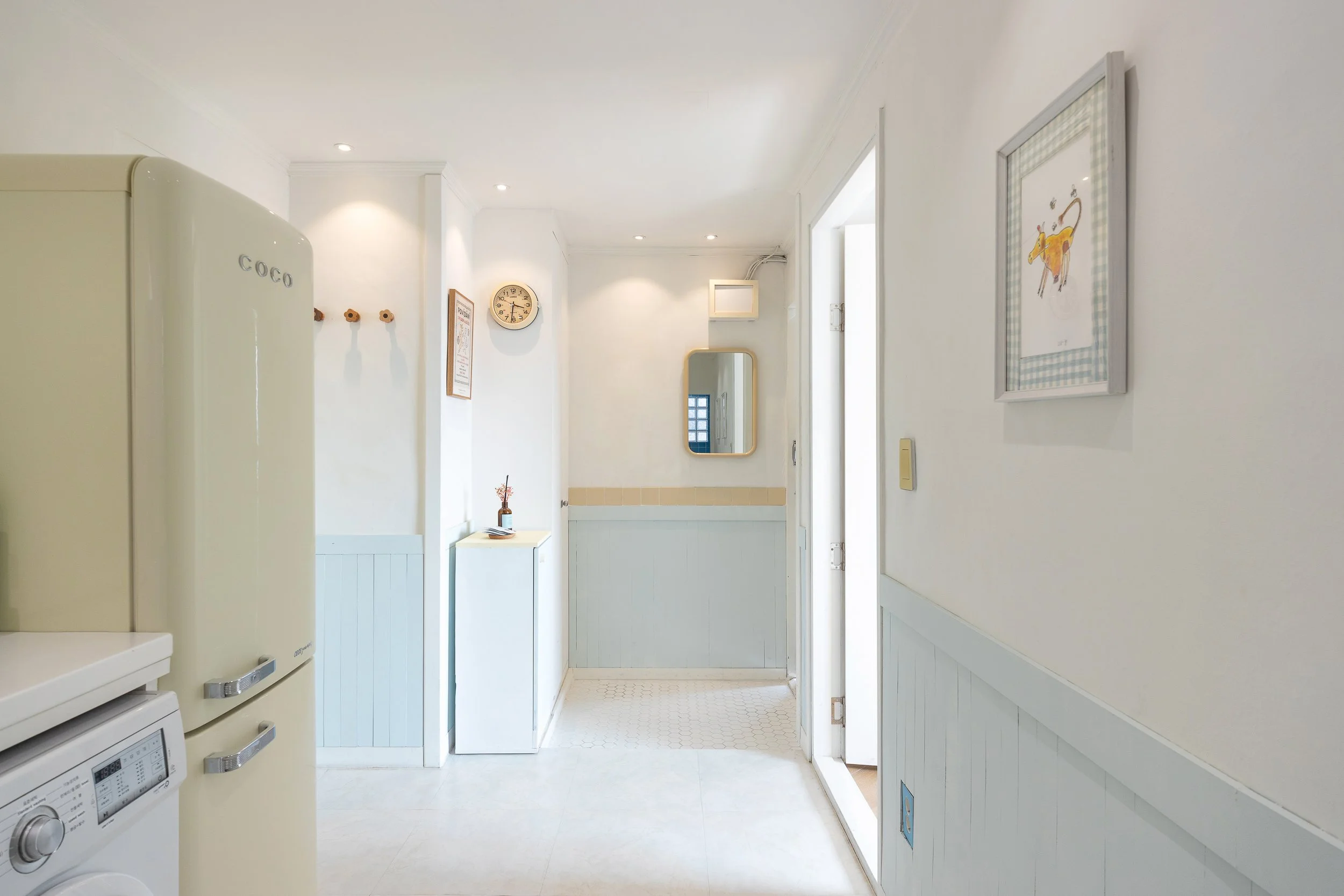 Bright, white-walled laundry area with blue paneling, featuring a cream-colored vintage-style refrigerator and a washing machine. Decor includes framed art, a wall clock, a mirror, and small decorative items.