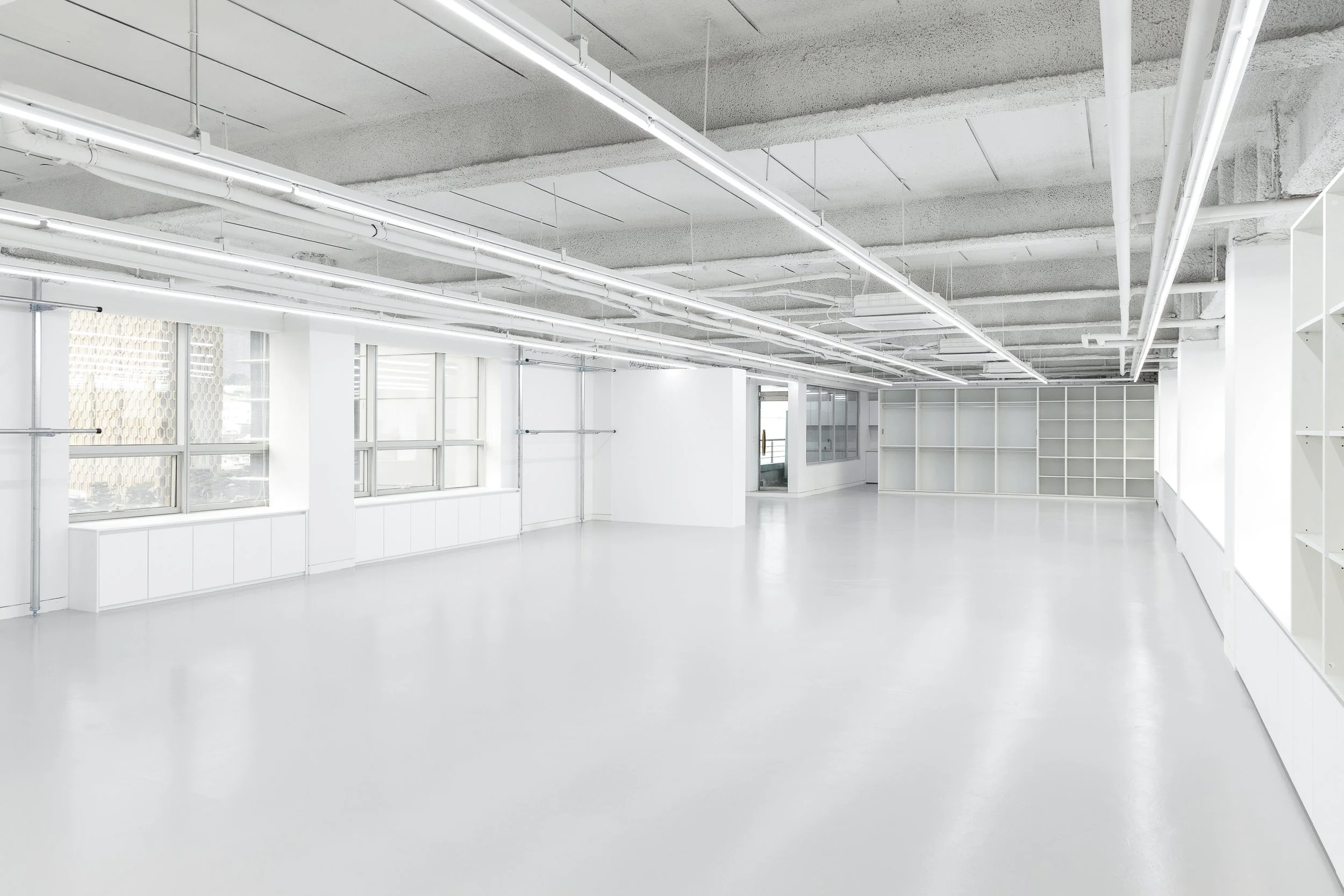 Empty, white-walled modern room with large windows and built-in shelves, featuring an industrial ceiling with exposed pipes and bright, natural lighting.