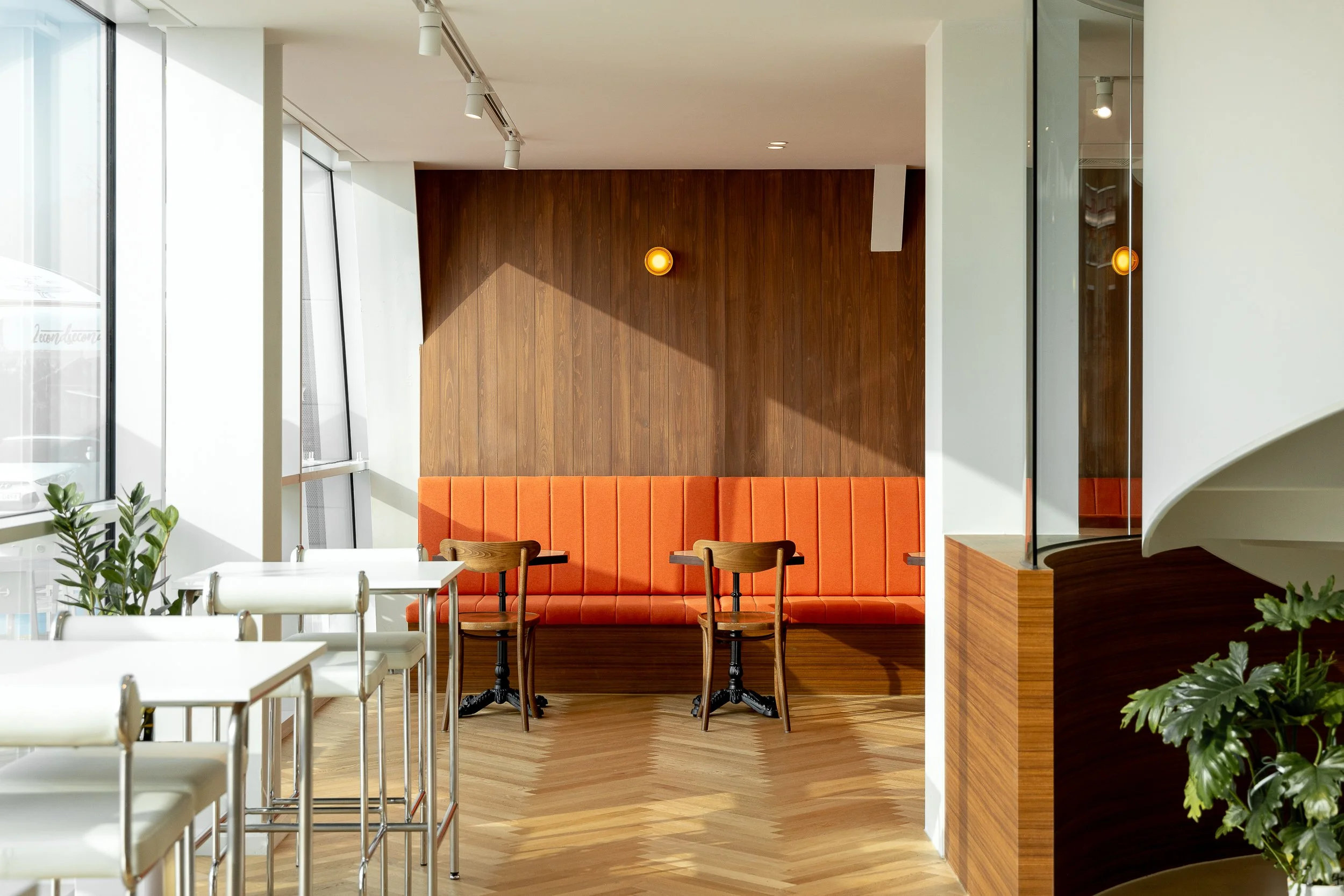 Modern cafe interior with white tables and chairs, orange upholstered banquette seating against a wooden paneled wall, and potted plants, illuminated by natural light from large windows.