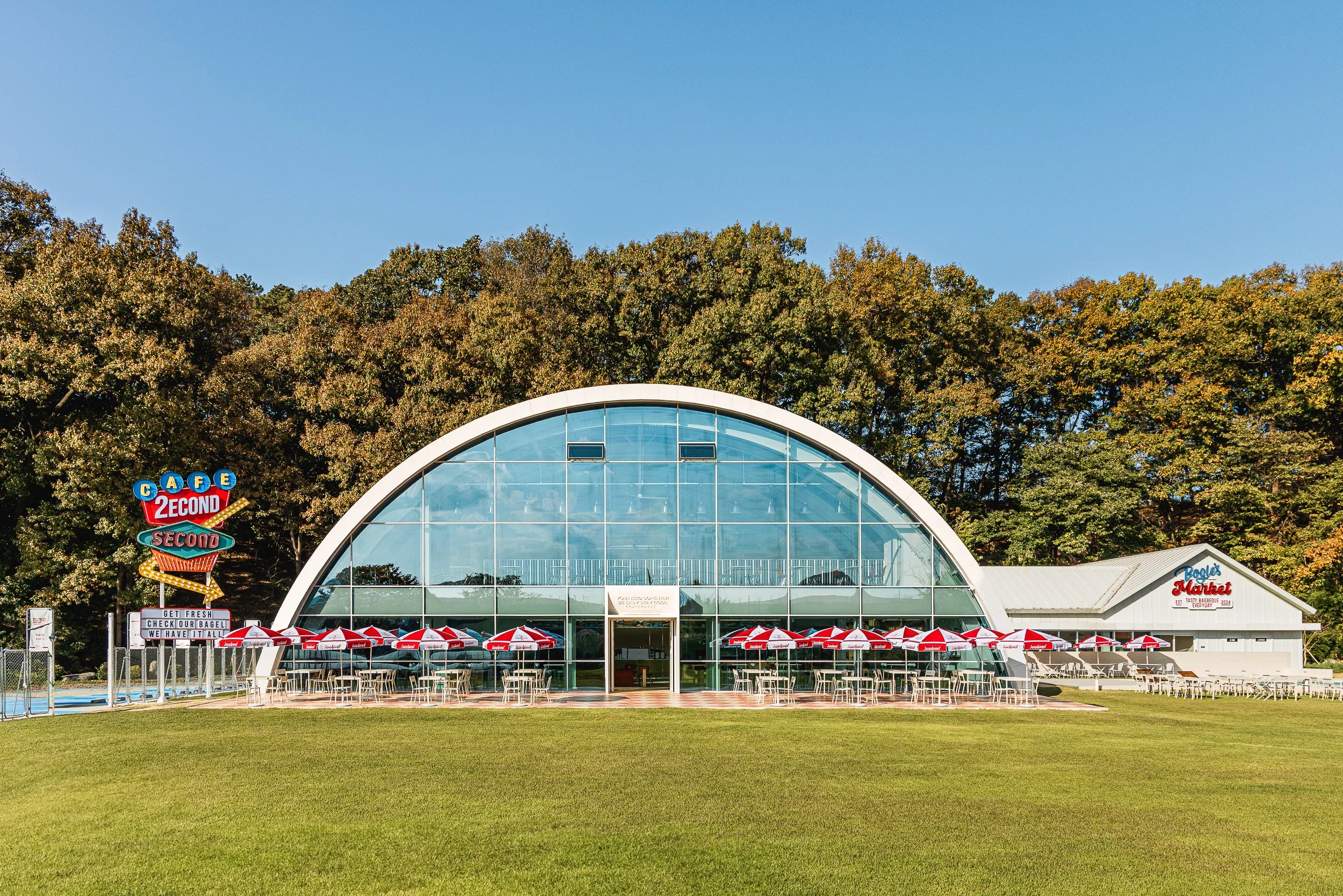 A large, glass-domed building with a white roof and multiple windows, surrounded by outdoor tables with red and white umbrellas, and a sign for a breakfast and lunch restaurant named 'Baylor's Market.' Behind the building, there is a forested area wi