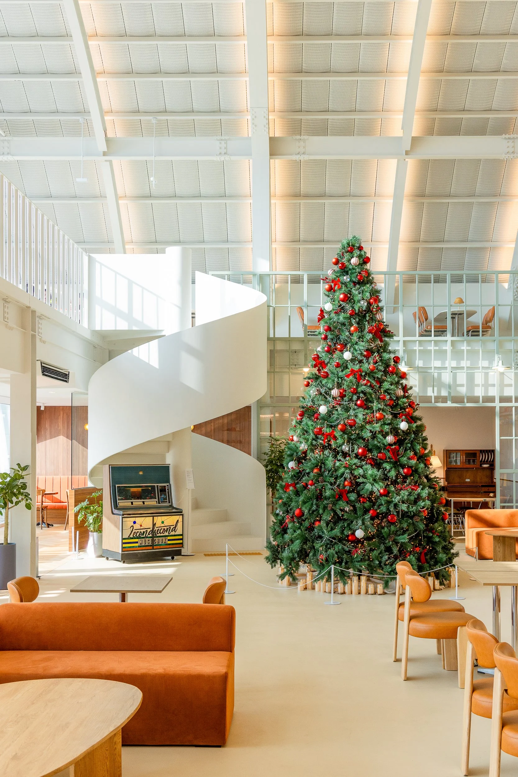 A large decorated Christmas tree in a modern, bright indoor lounge area with seating, a spiral staircase, and a glass-walled upper level.