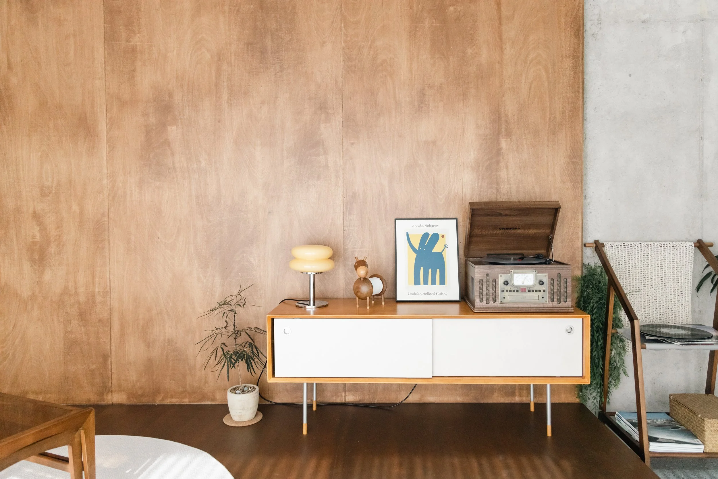 Living room decor with a white and wood sideboard, a potted plant, a vintage radio, framed artwork, and a chair with magazines on a shelf against a wood-paneled and concrete wall.