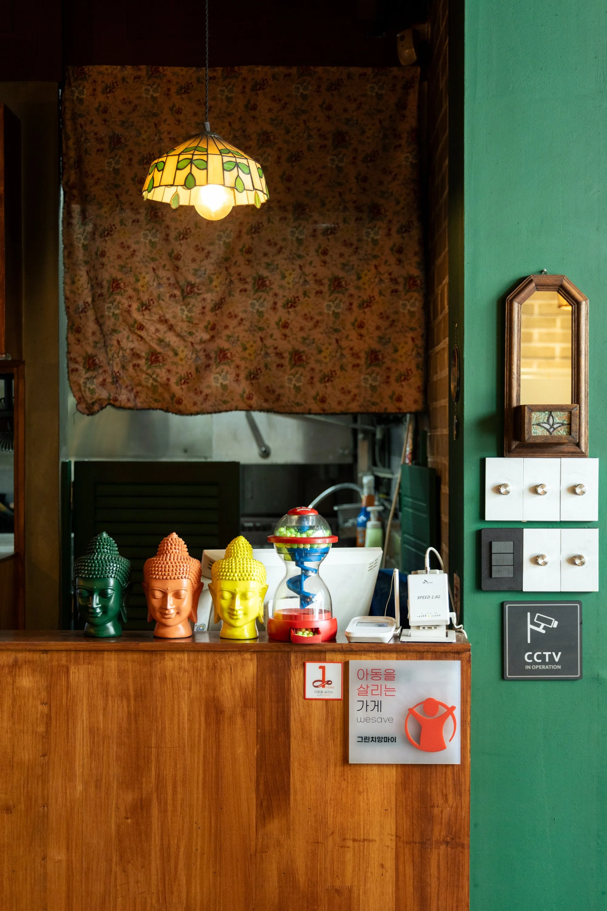 Reception desk with colorful Buddha head sculptures and a children's toy, inside a cafe or store with green wall, CCTV sign, and a floral curtain.