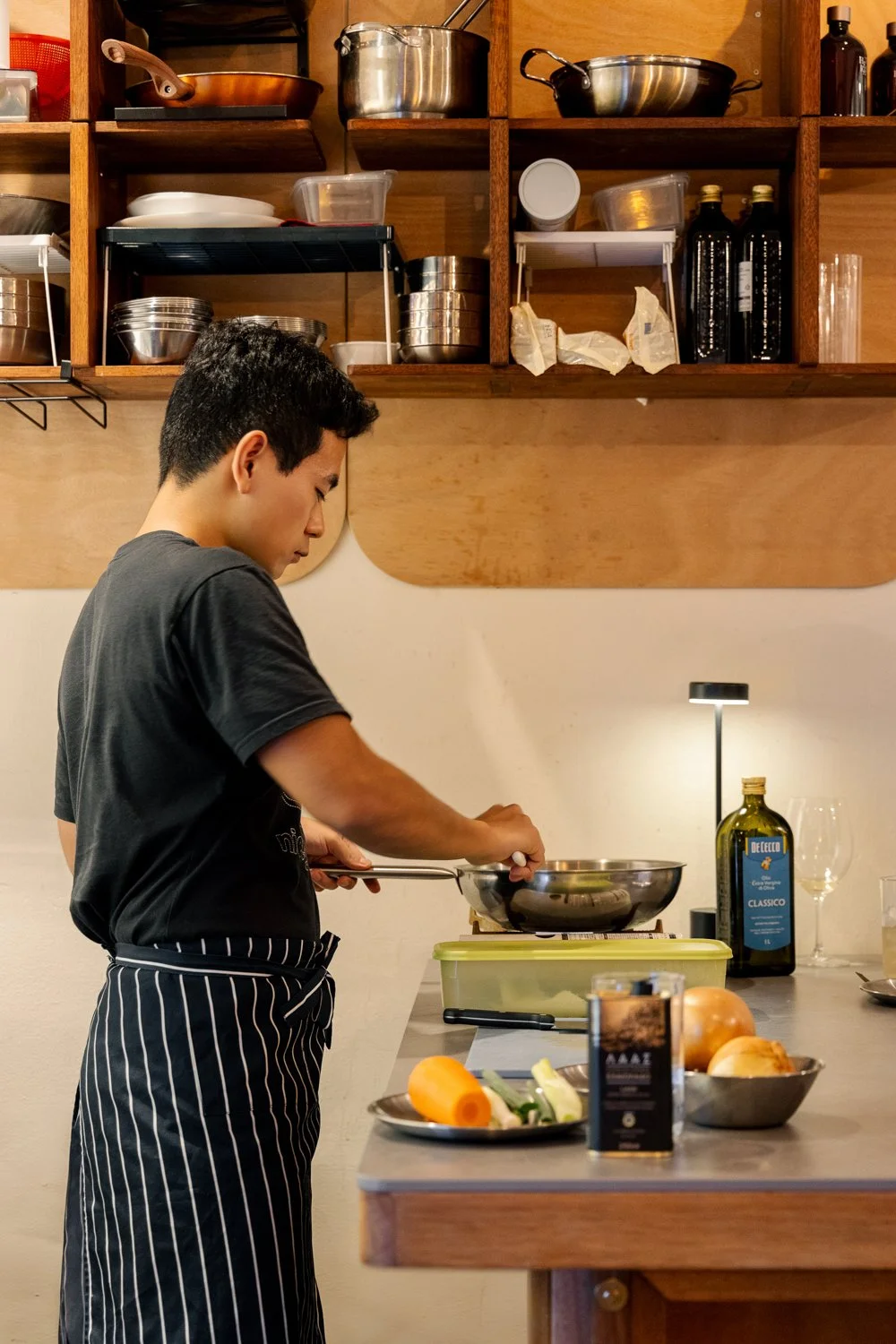 A young man wearing a black t-shirt and striped apron cooking on a stove in a kitchen with wooden shelves filled with pots, pans, and bottles.
