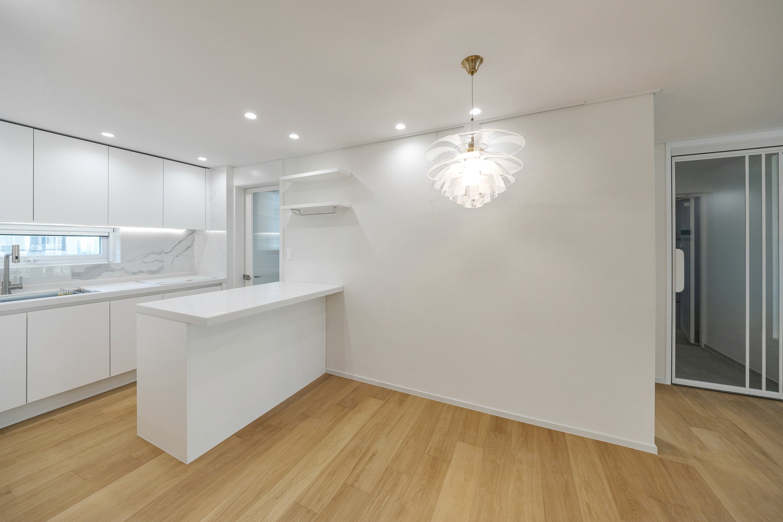 Bright modern kitchen with white cabinets, marble backsplash, and a wood floor, illuminated by recessed ceiling lights and a hanging light fixture.