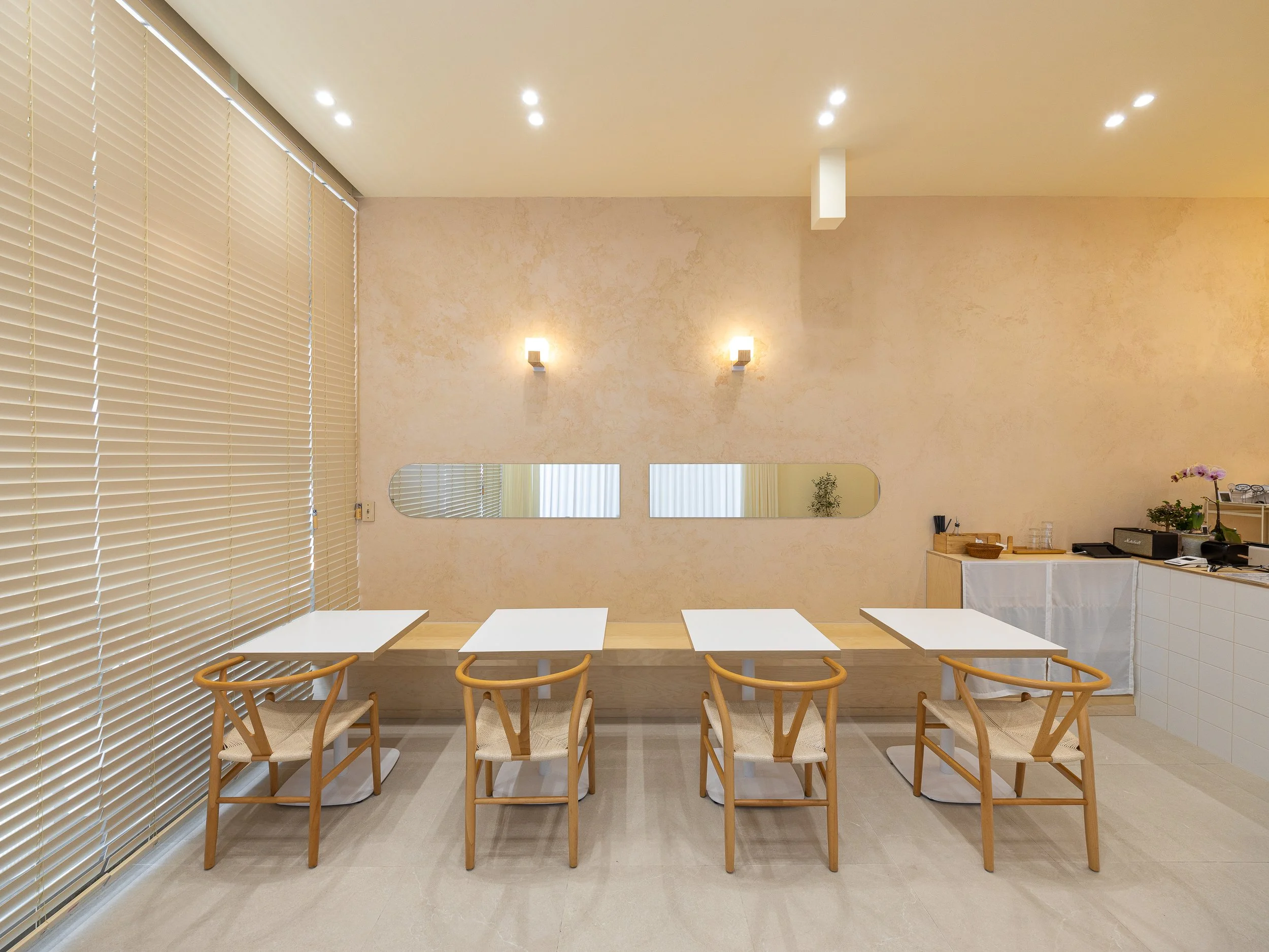Minimalist interior with four wooden chairs and white tables, beige textured wall with mirror, blinds covering large window, and small counter with plants and utensils.