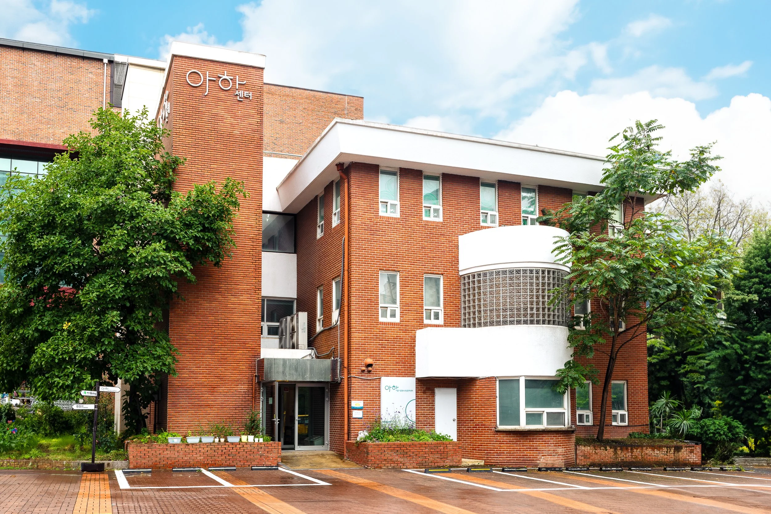 Red brick building with several windows and a curved white balcony, surrounded by green trees, under a partly cloudy sky, with a brick-paved parking area in front.