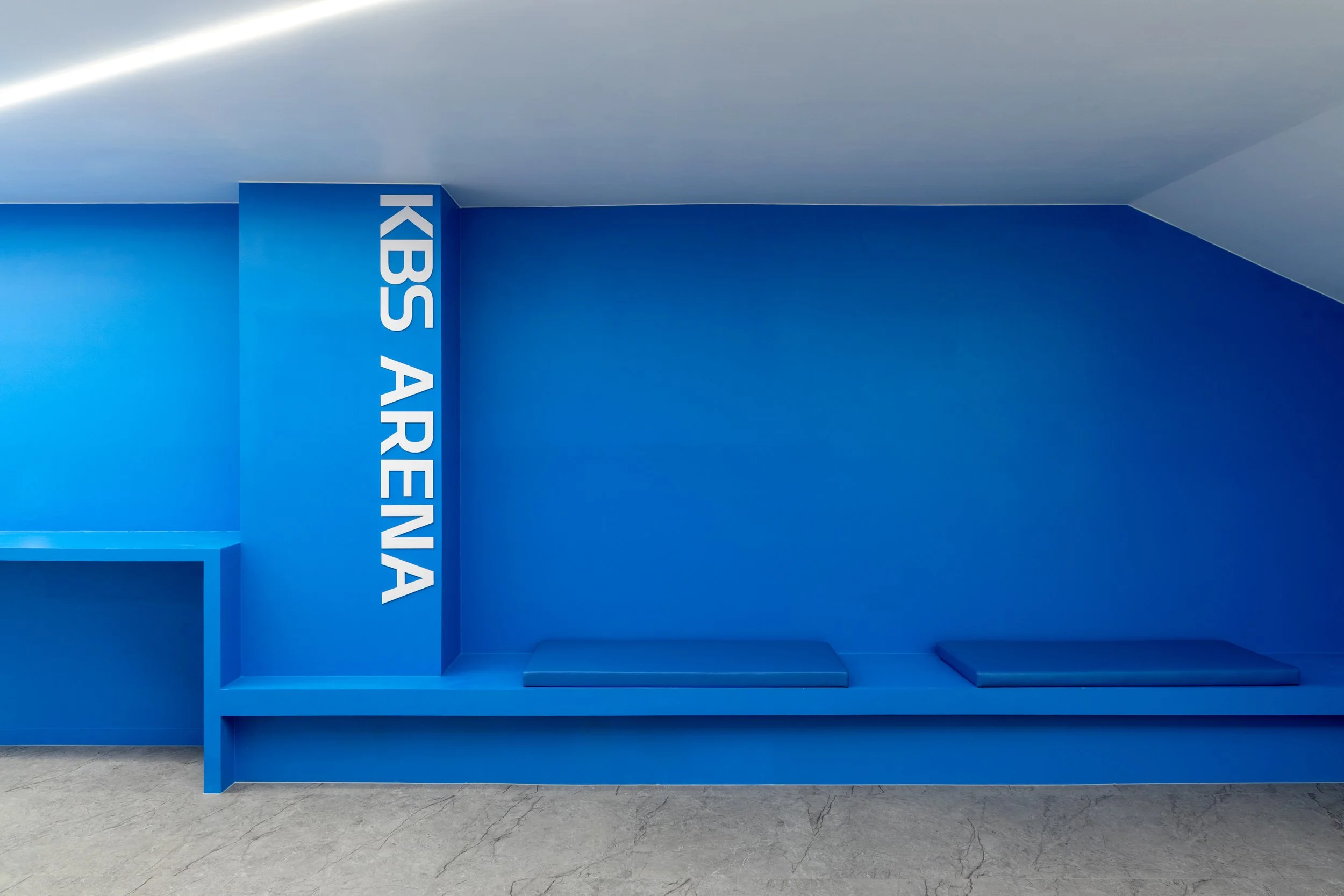 Blue reception area with a bench and white text that reads "KBS ARENA" on the wall.