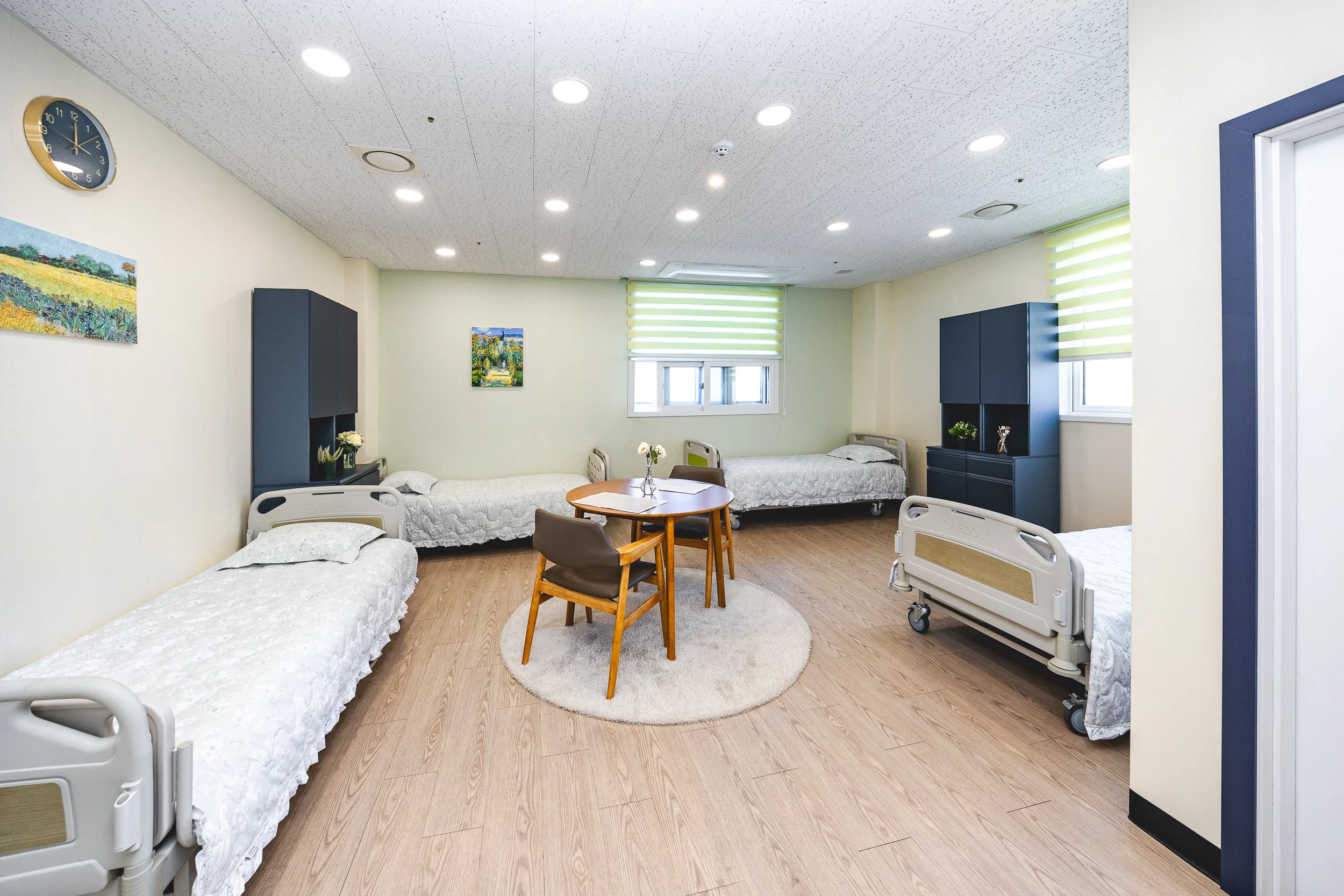 A hospital ward with four beds, two on each side, and a small round rug in the center with a wooden table and chairs. The walls are decorated with paintings, and there are windows with green striped blinds. The room has a ceiling with multiple recess