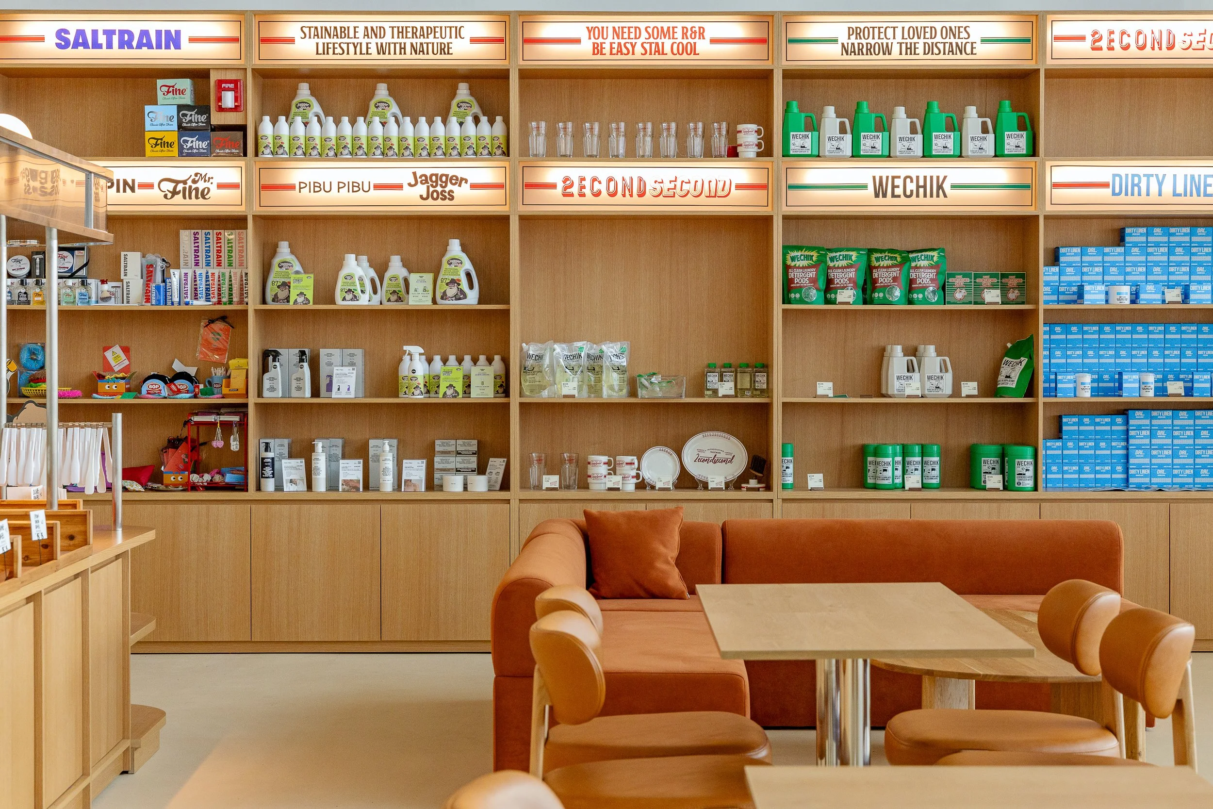 A retail store with wooden shelves displaying cleaning and household products, with signs overhead and a seating area with wooden tables and chairs in the foreground.