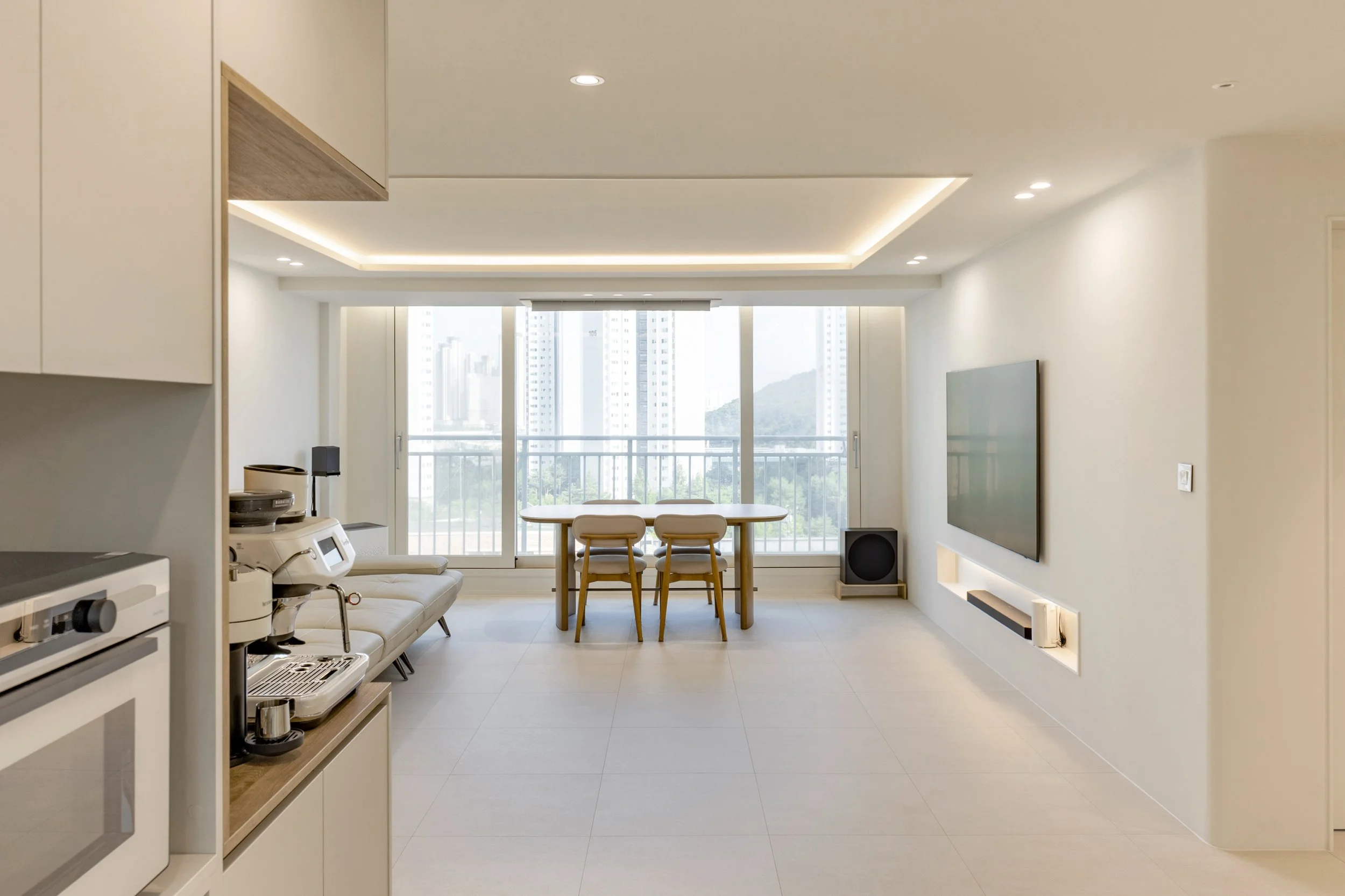 A modern, minimalist living room with a large window balcony view of city high-rises and hills; includes a dining table with three chairs, a wall-mounted TV, a sofa, and kitchen appliances.