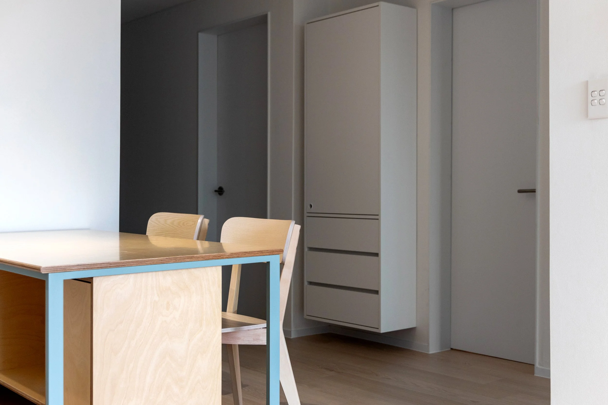 A modern dining area with a wooden table and matching wooden chairs with light-colored seats. In the background, there are white built-in storage cabinets and a door, with a light-colored wooden floor.