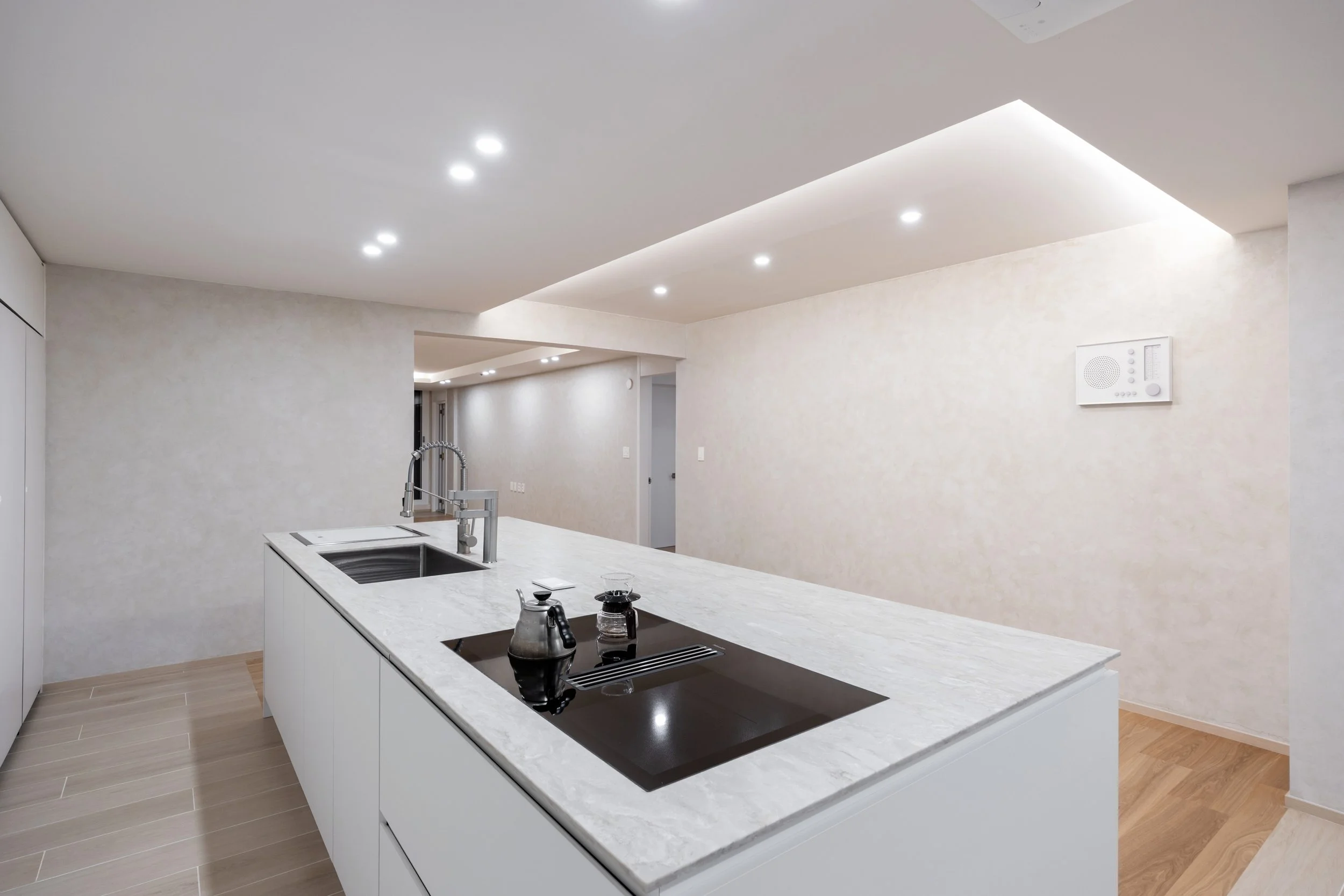 Modern kitchen with white marble island, induction stove, sink, and minimalistic decor; recessed ceiling lights and neutral wall color.
